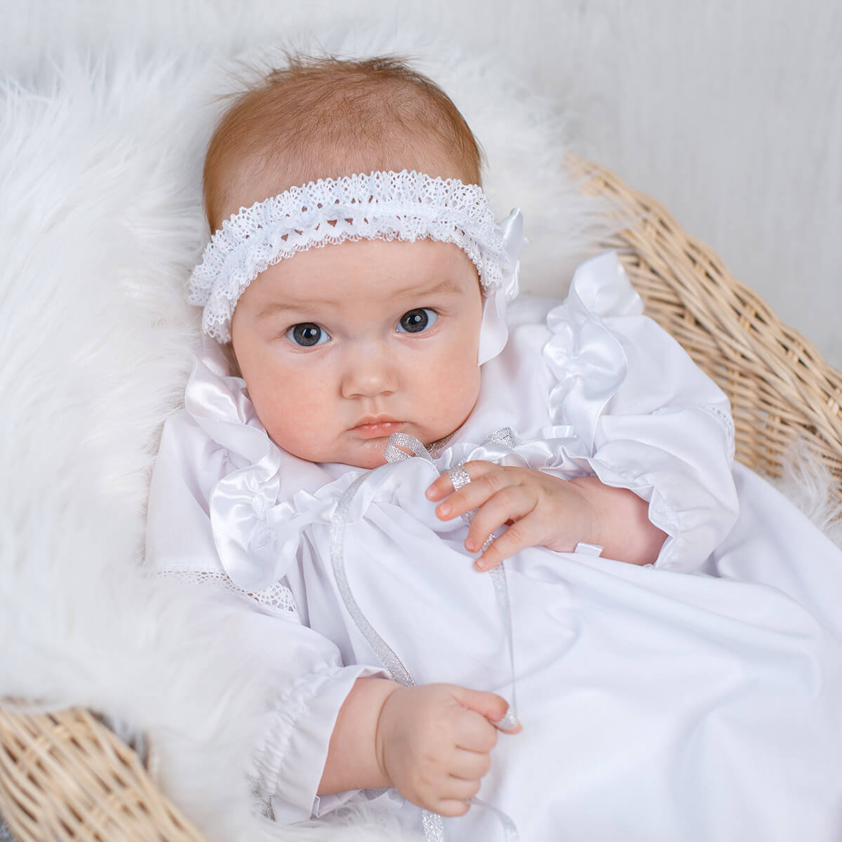 Baby in a white outfit with athe baby headband, lying on a white surface.
