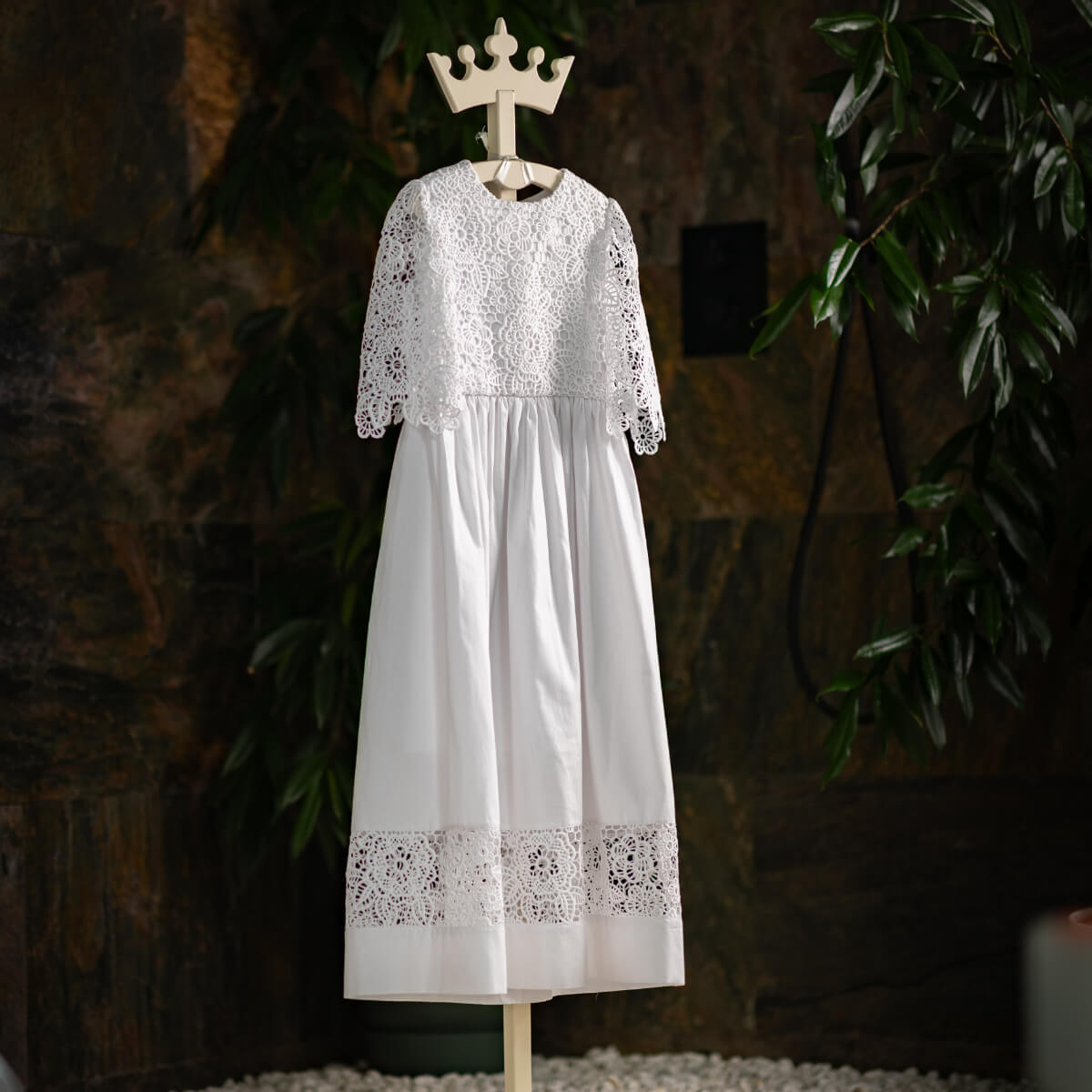 First Communion Cotton Dress on a wooden hanger against a dark background with plants.