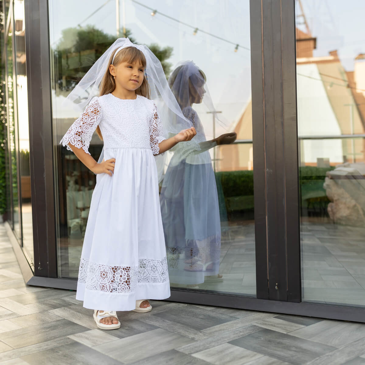 Young girl in a white dress with First Communion Veil standing in front of a glass door.