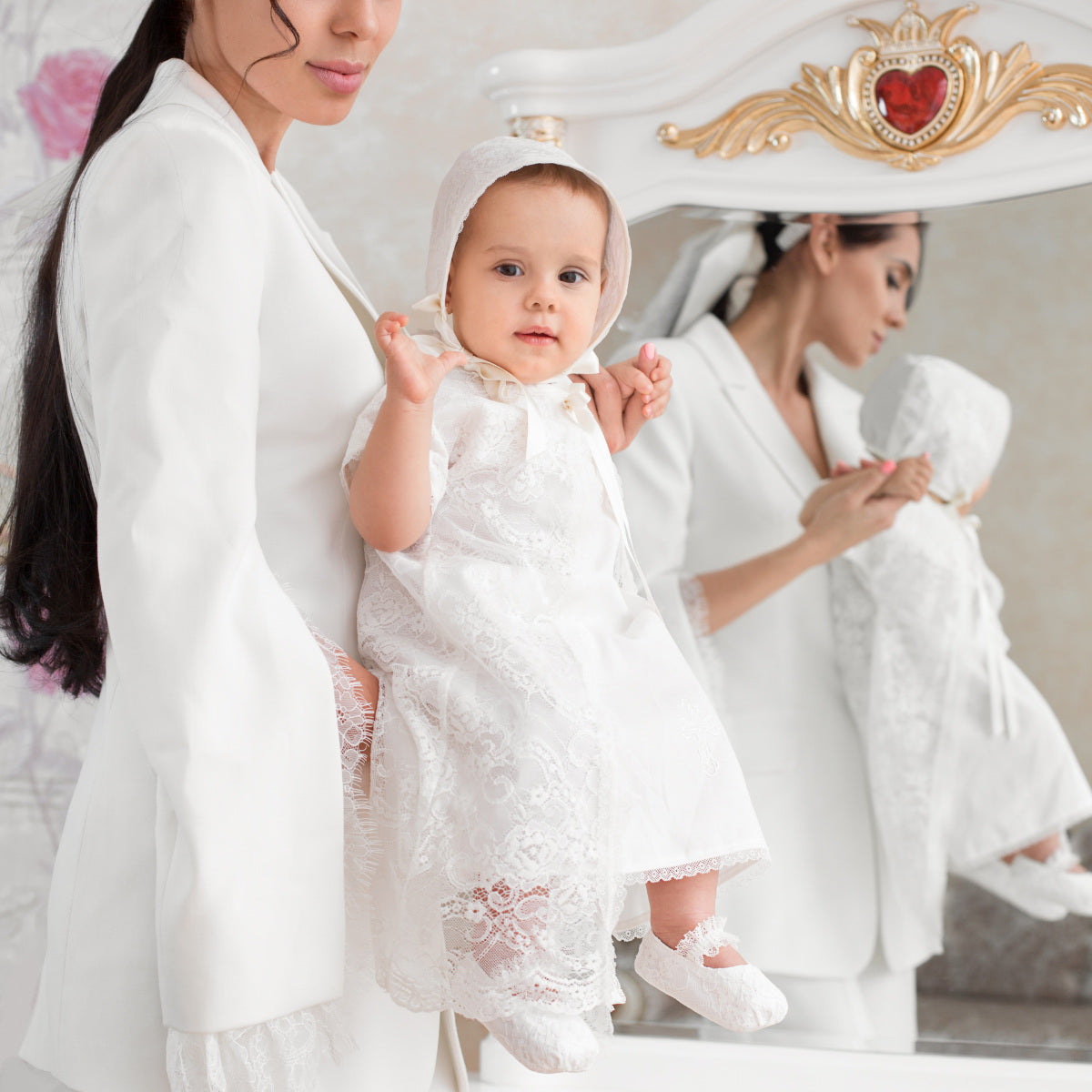 Woman holding a baby in a Girls Classic Baptism Gown with lace details, standing in front of a mirror.