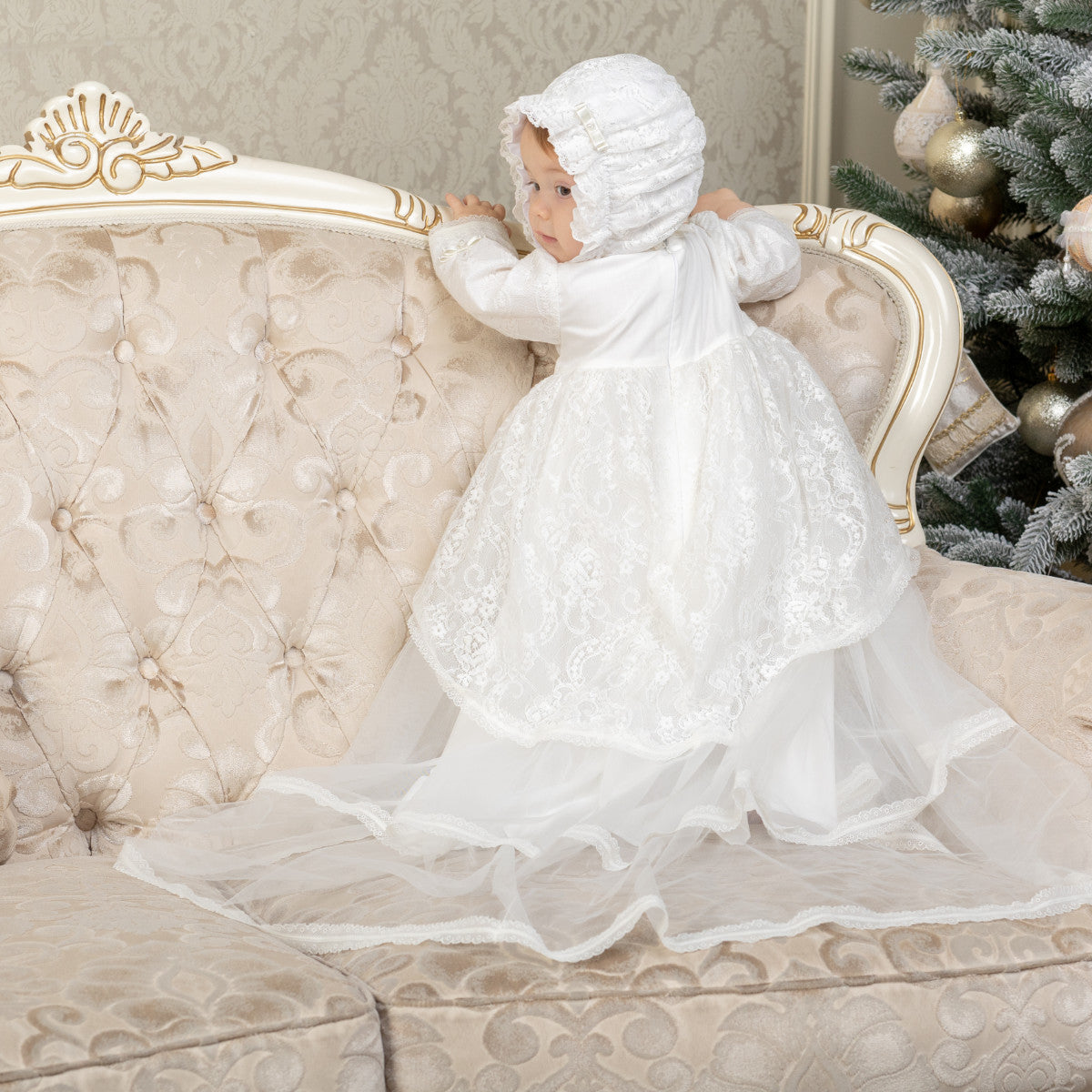 Child in a Christening Gown for Girls and bonnet sitting on an ornate couch with a decorated Christmas tree in the background.