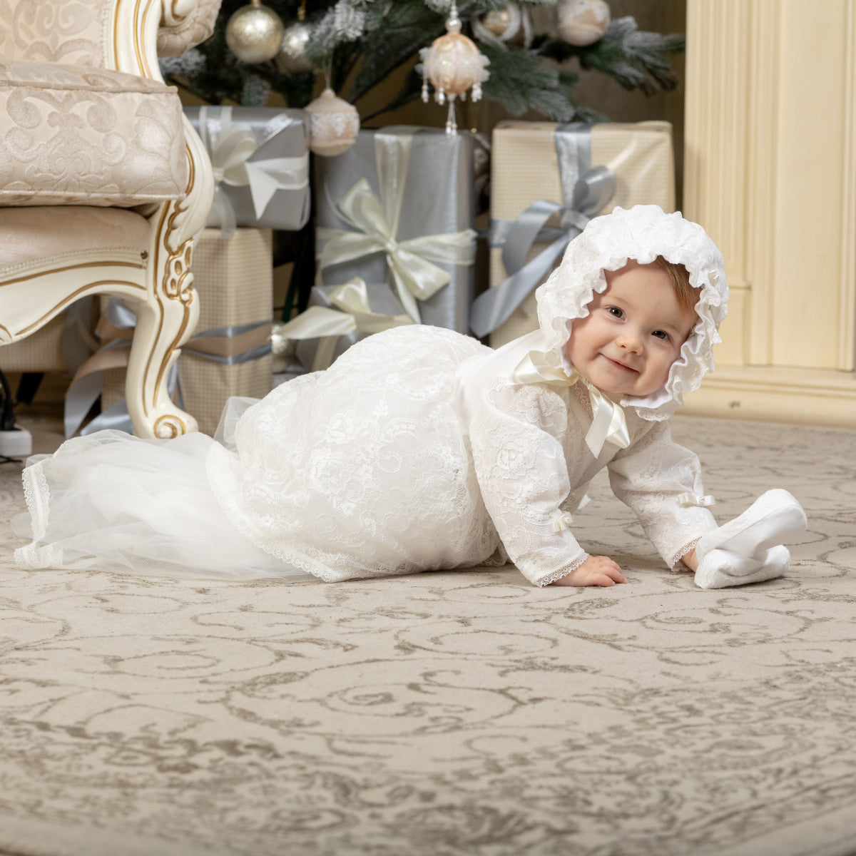Baby in a Christening Gown for Girls and bonnet crawling on a patterned rug with Christmas decorations in the background.