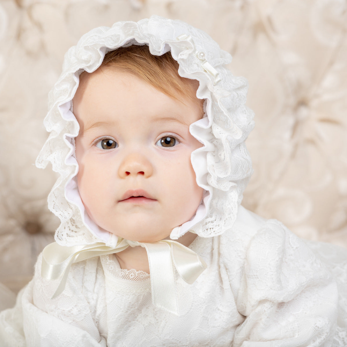 Baby wearing a lace christening bonnet with ruffles against a soft beige background