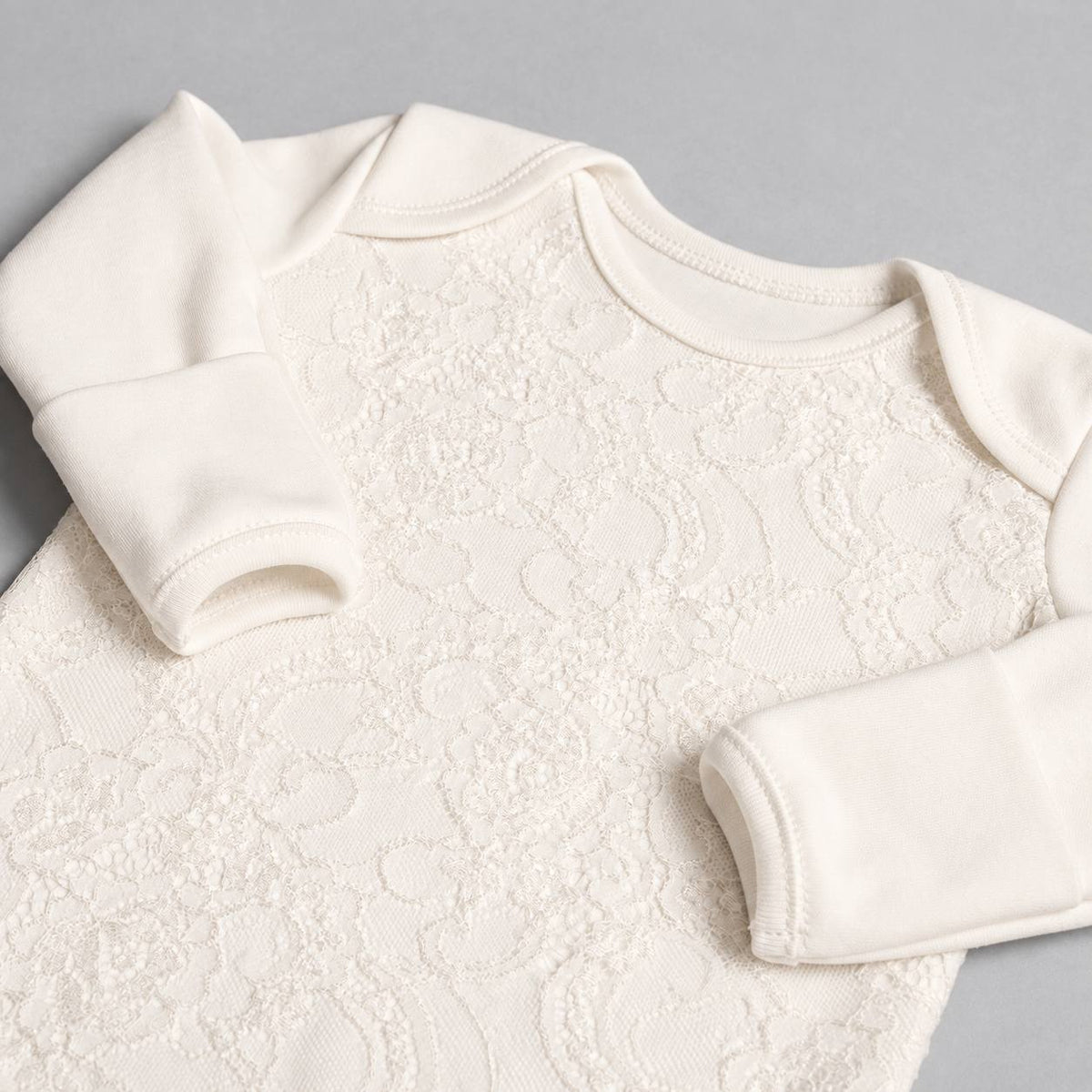 An off-white long-sleeve baby bodysuit featuring a textured floral lace pattern across the chest, laid flat on a neutral gray background.