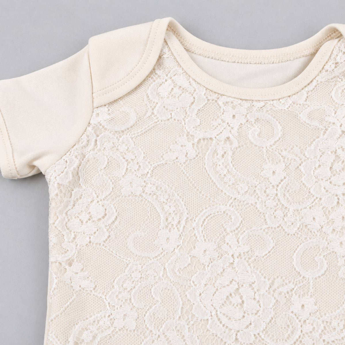 Close-up of a baby bodysuit featuring a cream-colored lace overlay on a soft beige knit base, with short sleeves and a ribbed crew neckline, shown on a neutral gray background.