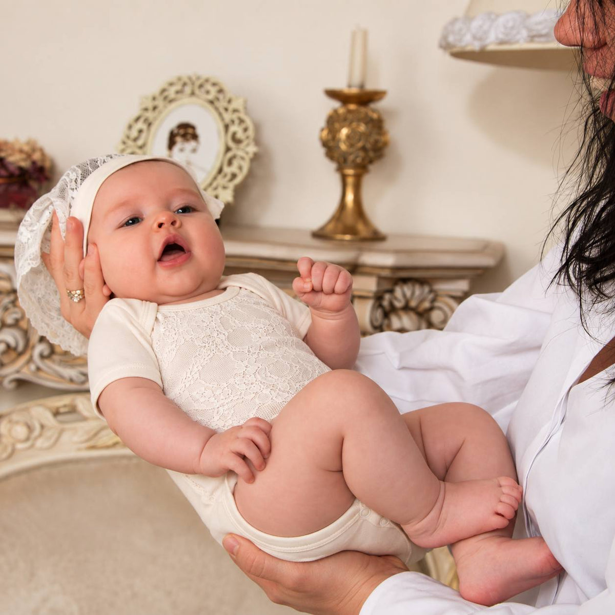 A baby wearing a cream lace short-sleeve bodysuit and matching bonnet is held by an adult in a white robe in an elegant indoor setting.