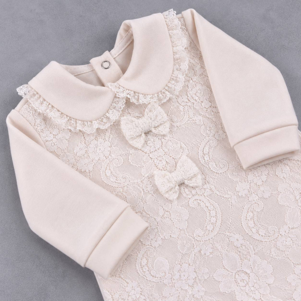 A close-up of a baby garment in a soft cream or off-white color, featuring a lace front panel with floral embroidery, a wide collar with ruffled trim, and two small bow details on the chest. The garment is laid flat on a neutral gray background.