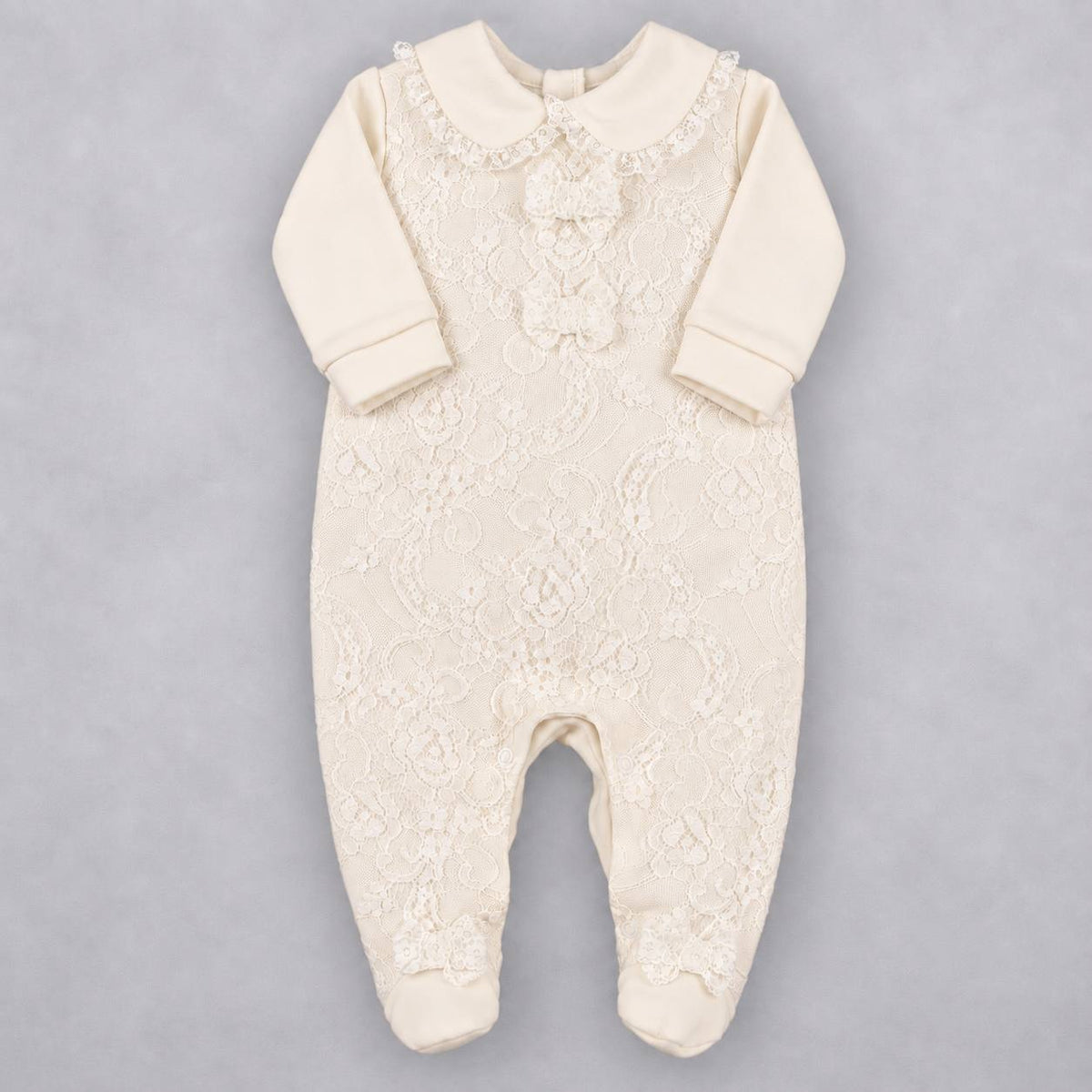 An off-white/cream baby footed sleeper with long sleeves, a lace-embroidered bodice, and a decorative ruffle collar, laid flat on a neutral gray background.