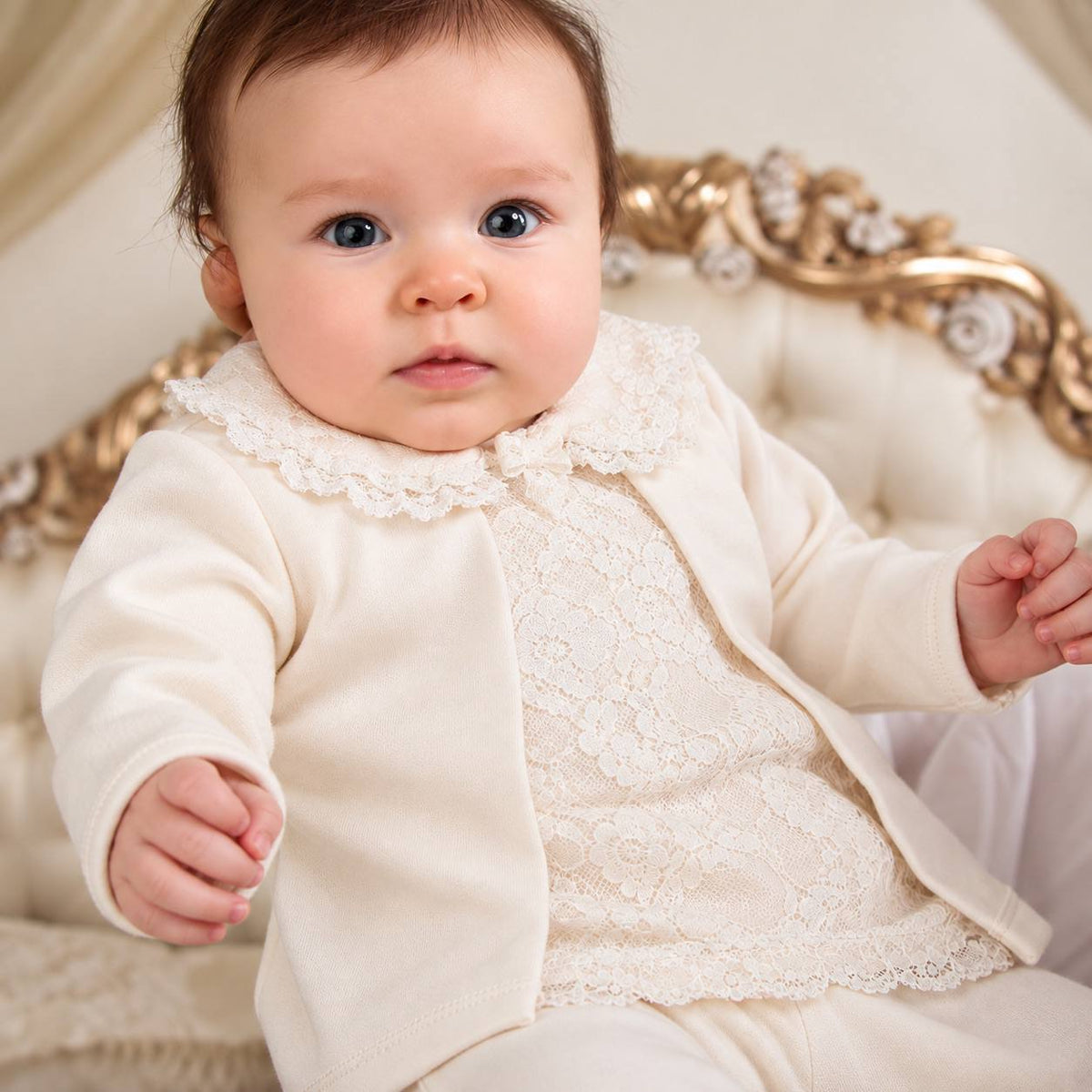 A baby wearing a cream-colored outfit with a lace-trimmed collar and matching cream cardigan, seated on an ornate gold and cream tufted chair.