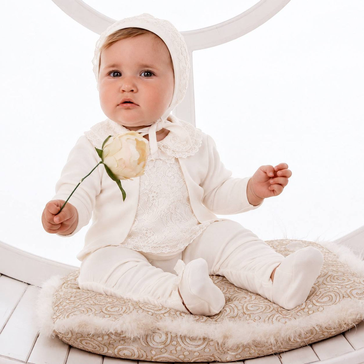 A baby wearing a cream-colored lace outfit with a bonnet sits on a patterned cushion, holding a pale rose.