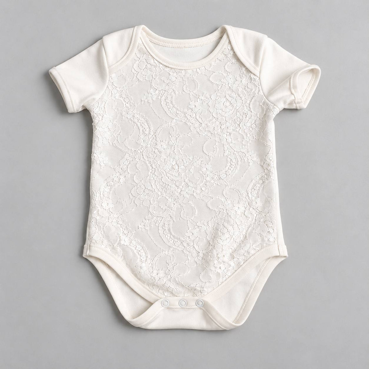 An off-white baby bodysuit with a short-sleeve design and an all-over floral lace overlay, laid flat on a neutral gray background.