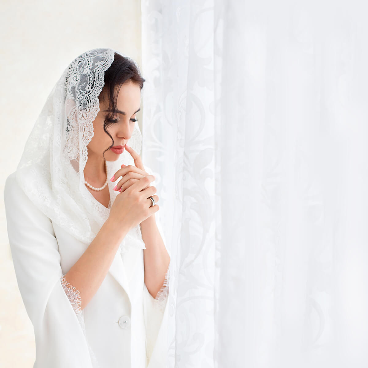 Woman wearing a Catholic Infinity Veil with lace details, standing behind a white curtain.