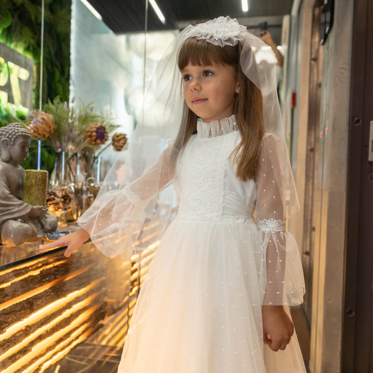 Young girl in a first communion dress with a veil standing indoors.