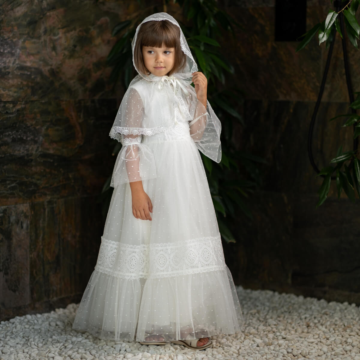 Young girl in a white lace first communion dress standing on pebbles with a dark background