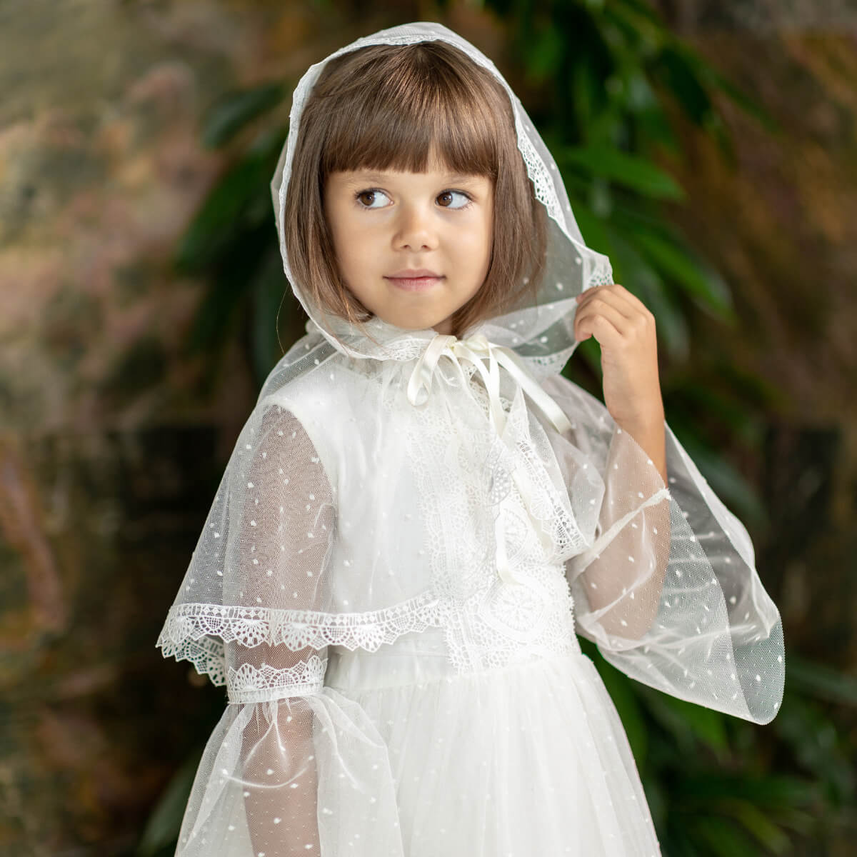 Young girl wearing a white lace dress with a First Communion Veil against a blurred green background