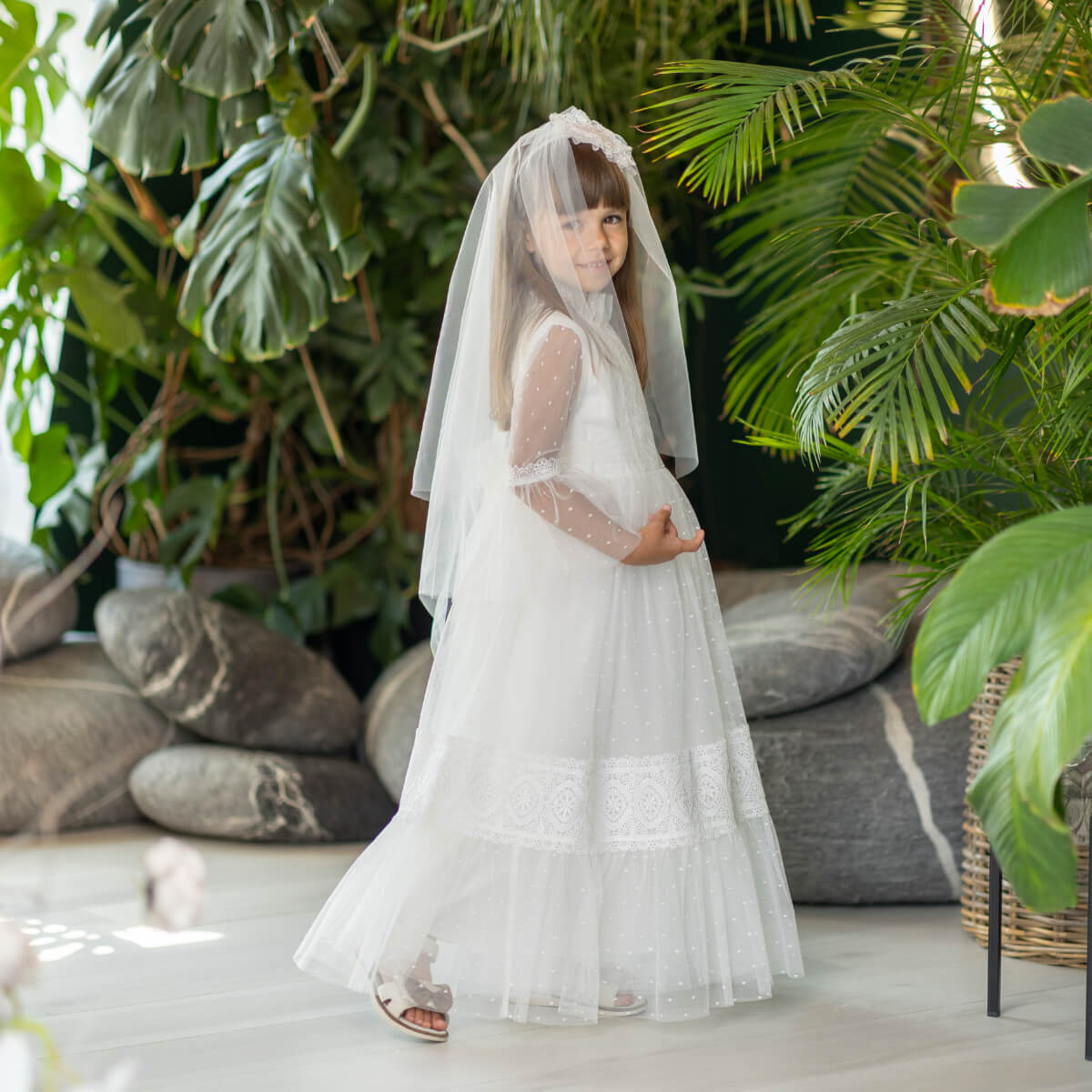 Child in a white dress with a First Communion Veil standing among green plants and stones.