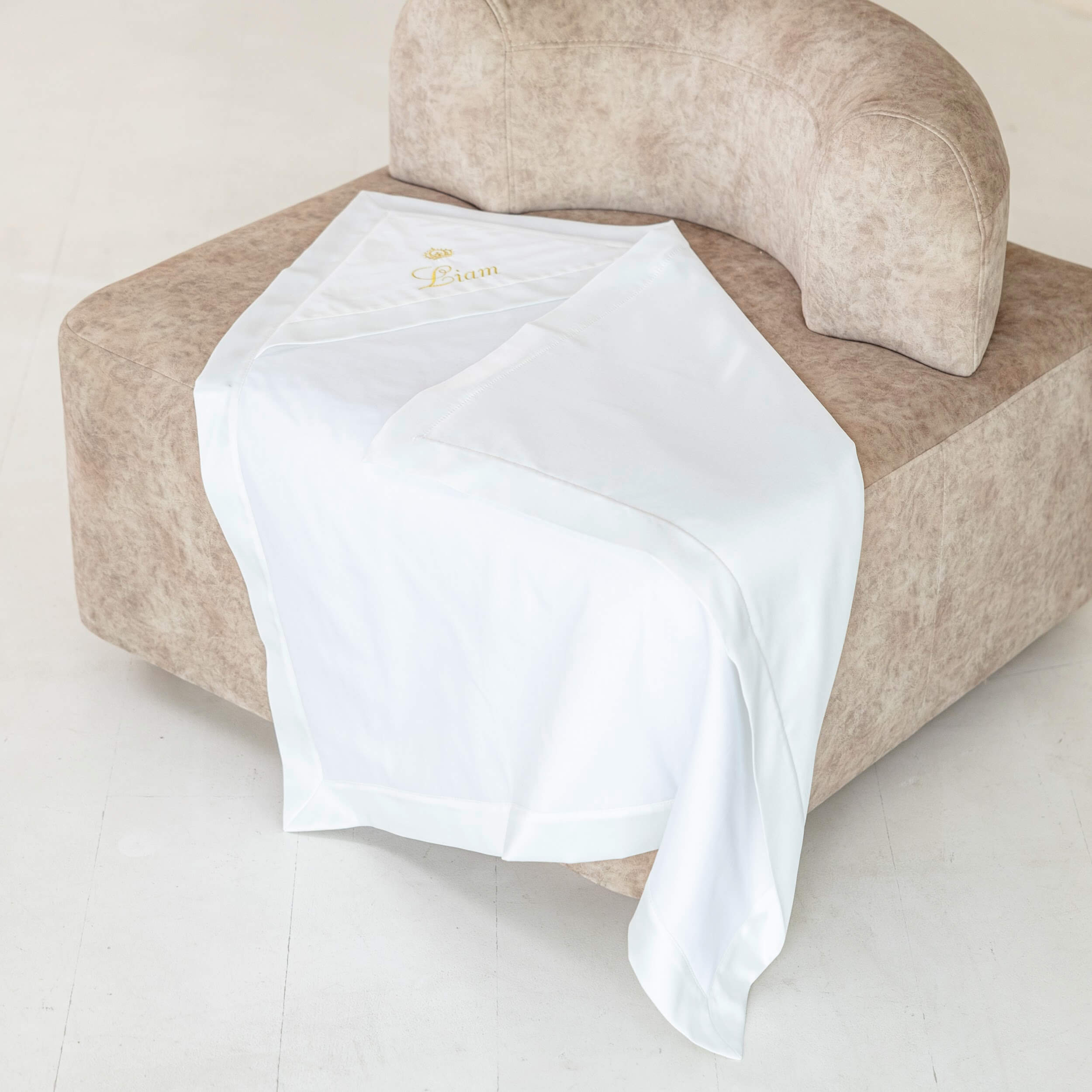 White baptism blanket draped over a beige armchair on a light wooden floor.