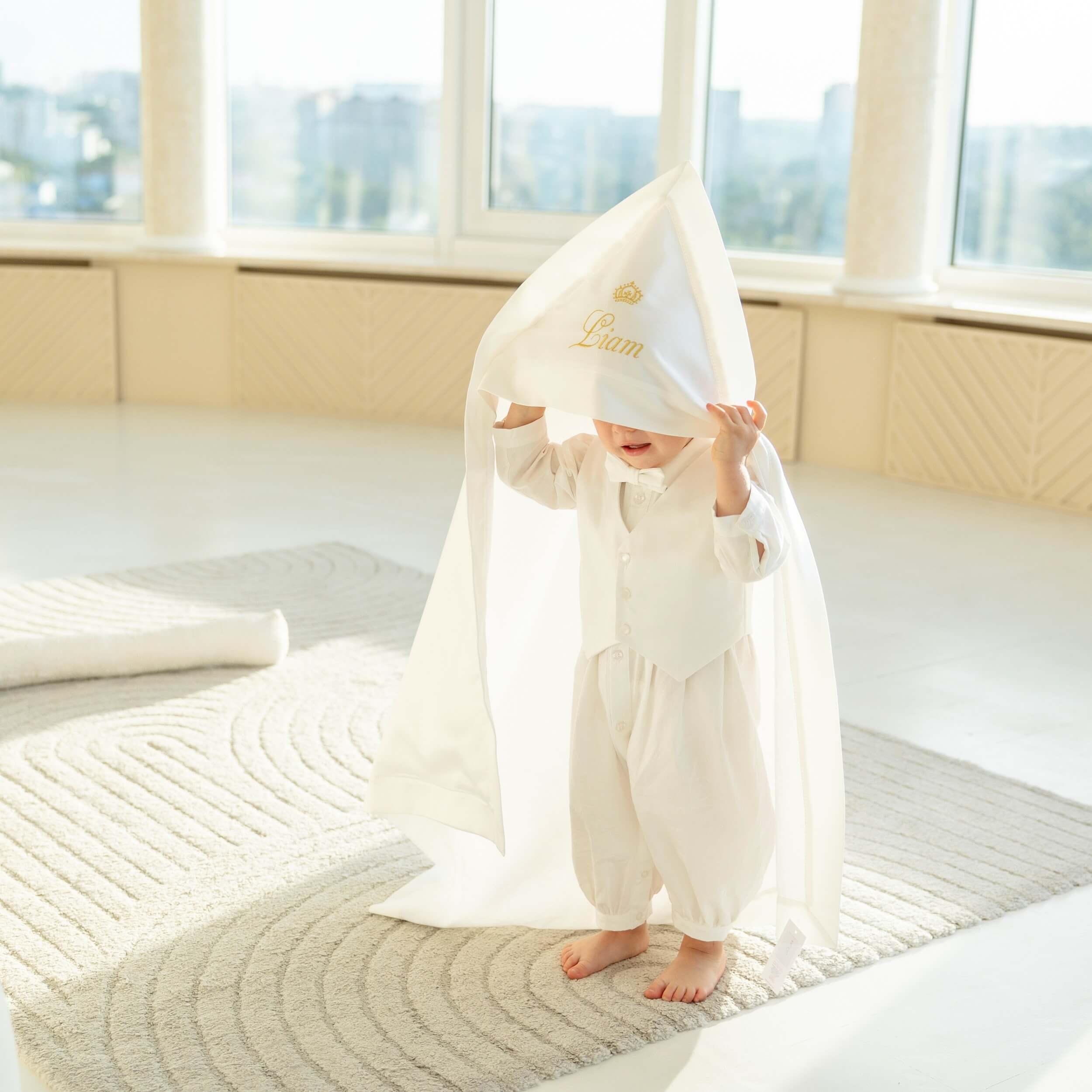 Baby in a white outfit standing on a rug with a large white hooded baptism blanket draped over them, in a bright room with large windows.