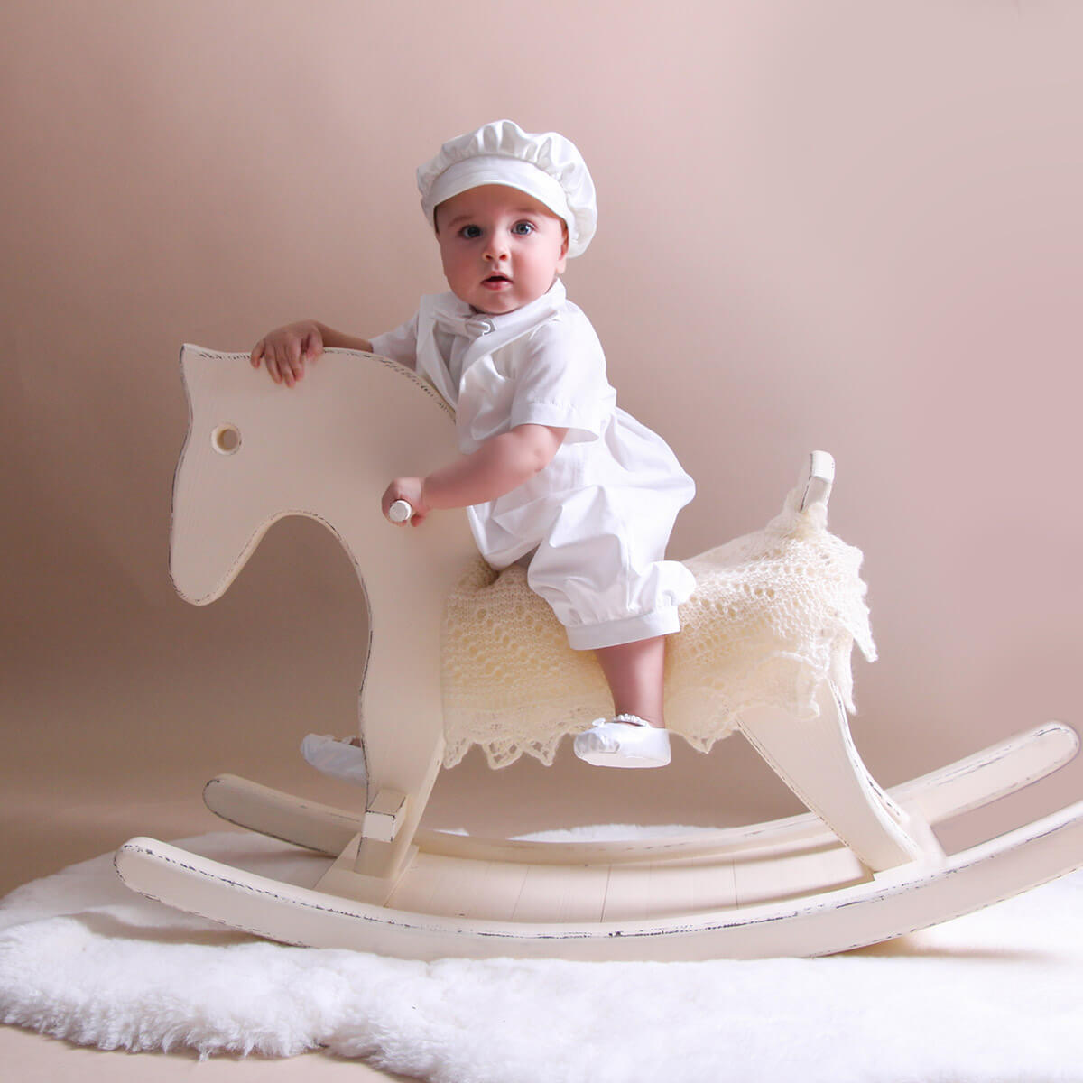 Baby in Baby Boy Baptism Romper sitting on a rocking horse against a beige background