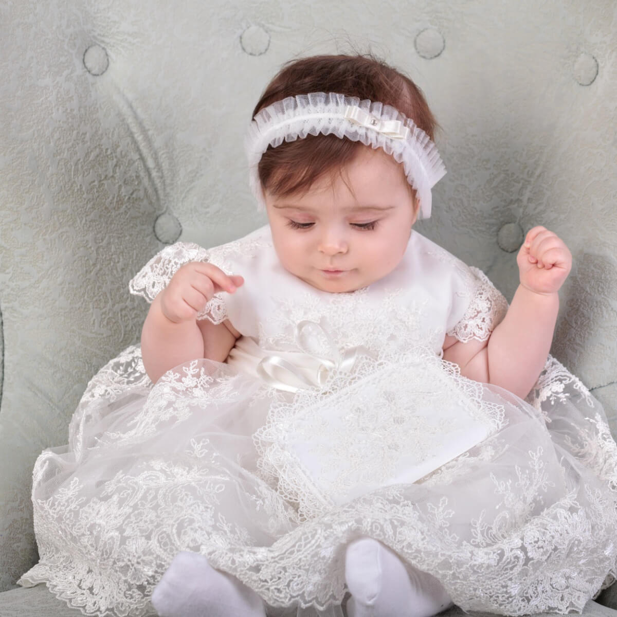 Baby in a white lace dresskeeping Baby First Curl Keepsake Box, sitting on a gray cushioned chair.