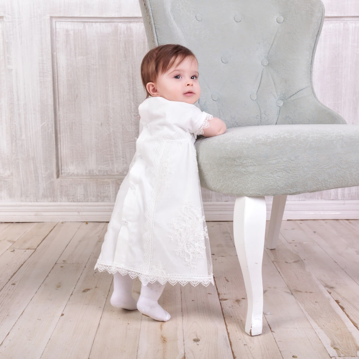 Baby in a. Girl Christening Gown standing next to a gray chair in a room with wooden floor and light-colored walls.