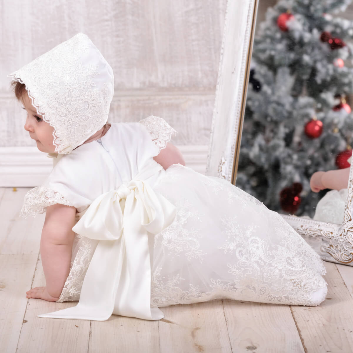 Baby in a Elegant Christening Dress sitting on a wooden floor with a Christmas tree in the background.