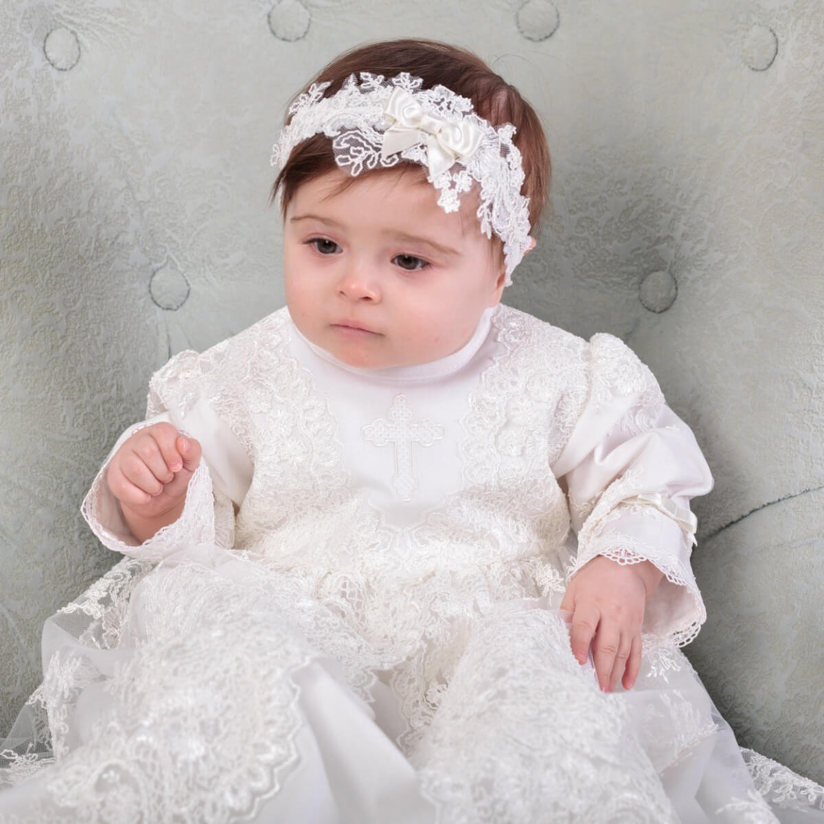 Baby in a white lace dress with the baby headband sitting on a gray cushion.