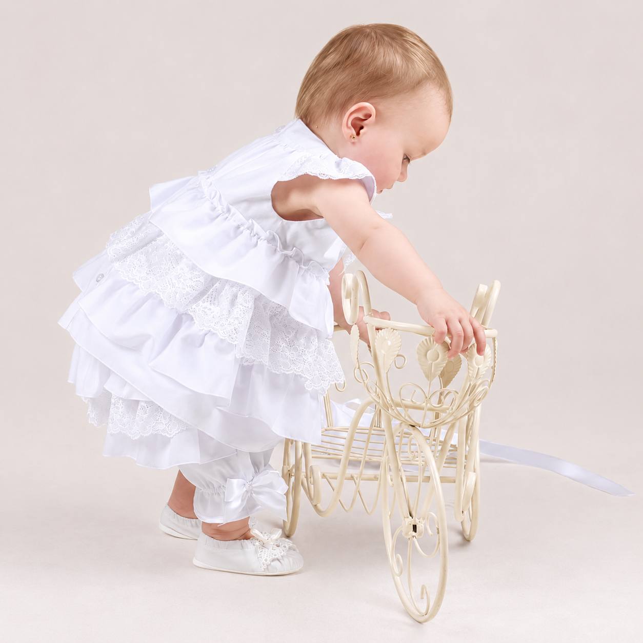 Baby in a white summer baptism dress playing with a small carriage on a plain background