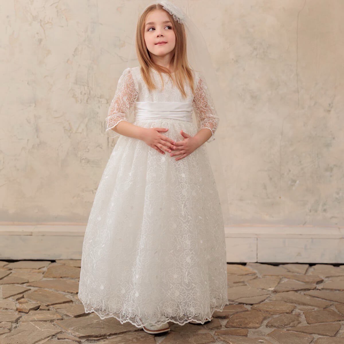 Young girl in First Communion Dress standing on a stone floor with a neutral wall background
