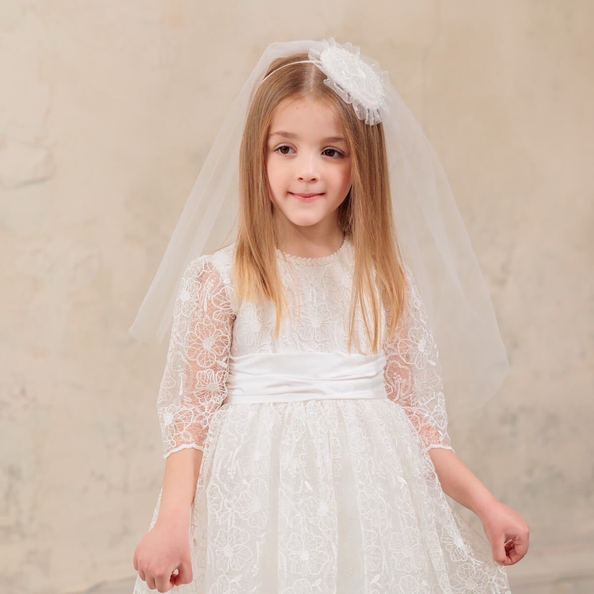 Young girl wearing a First Communion Dress with a matching veil against a beige background