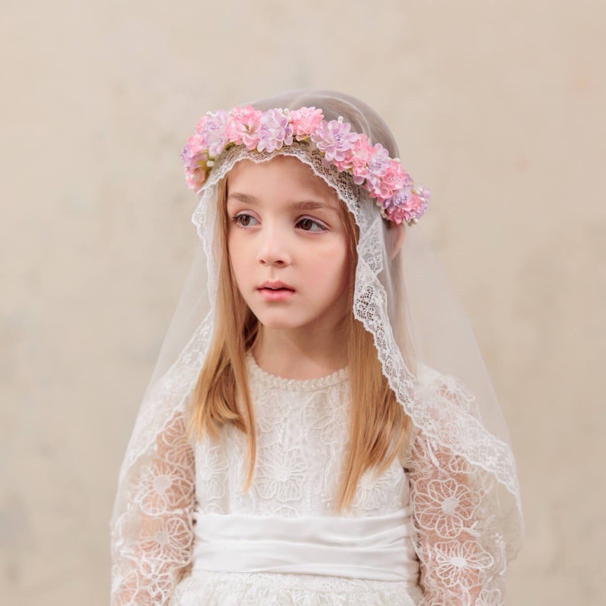 Young girl wearing a white lace dress and floral headband against a beige background