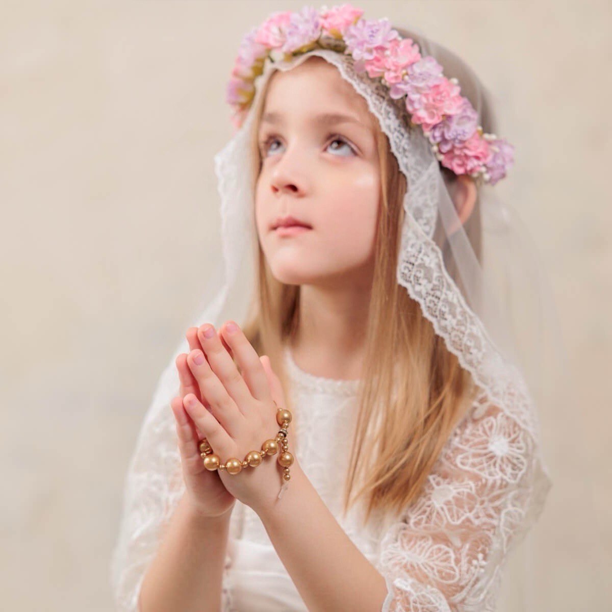 Young girl in a white lace veil with a floral headband, praying against a beige background