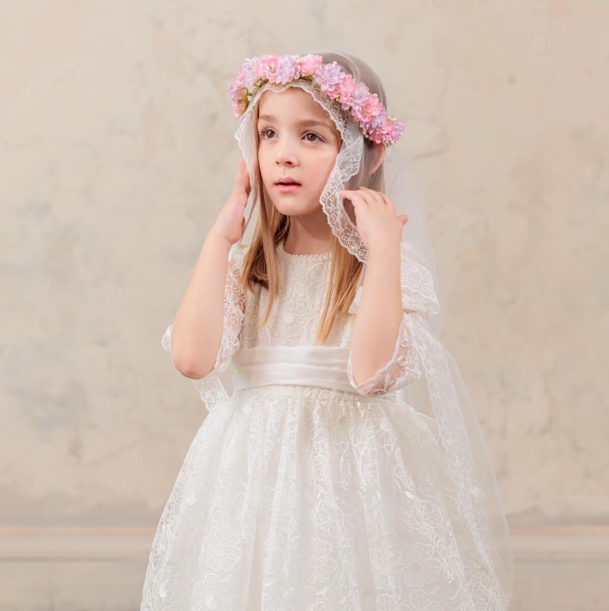 Young girl in a white lace dress with a floral headband against a beige background