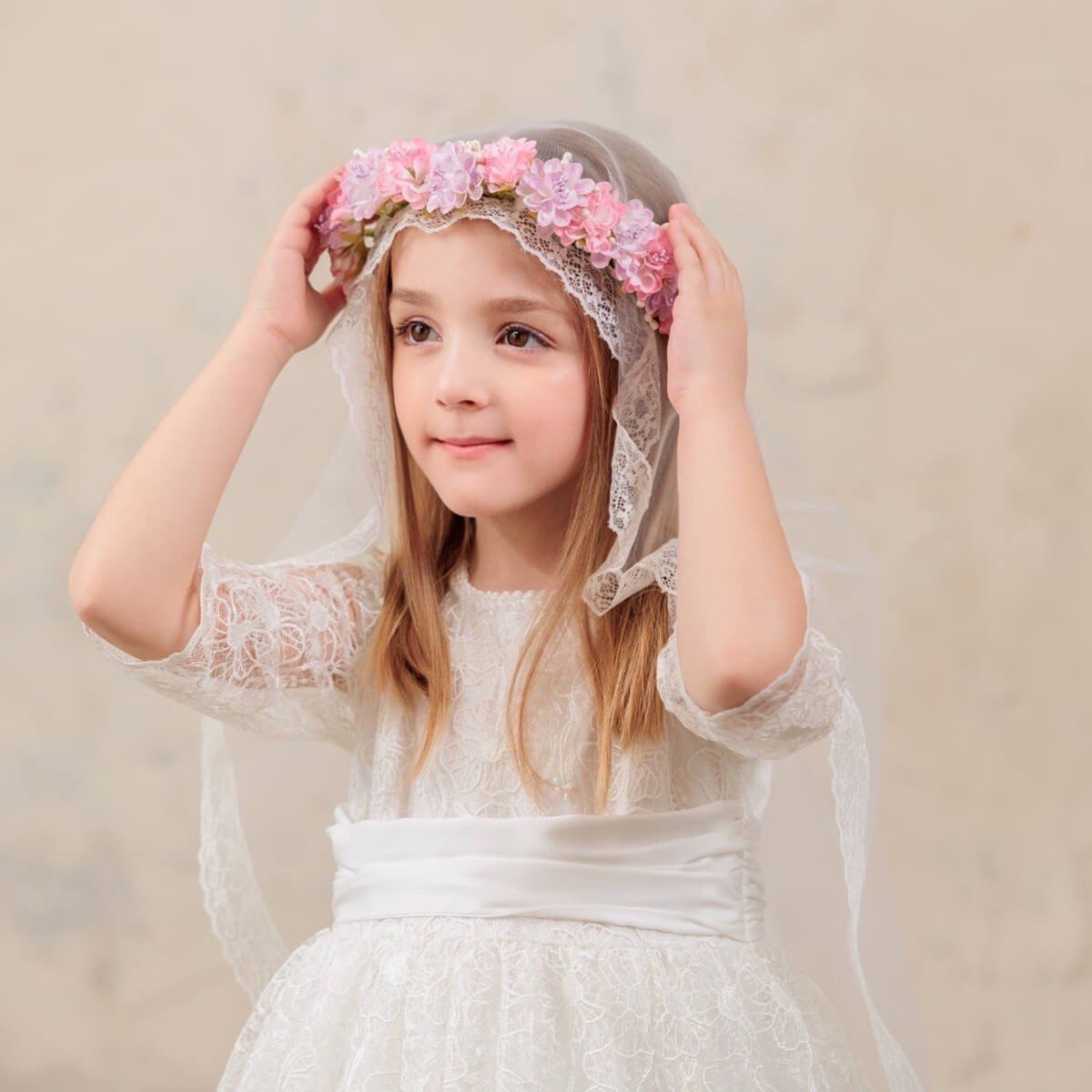 Young girl wearing a white communion dress with a floral headband against a beige background