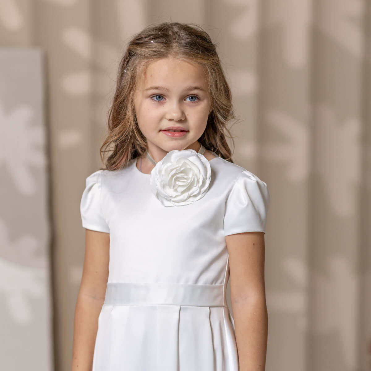 Young girl wearing a white simple communion dress with a large floral brooch against a neutral background