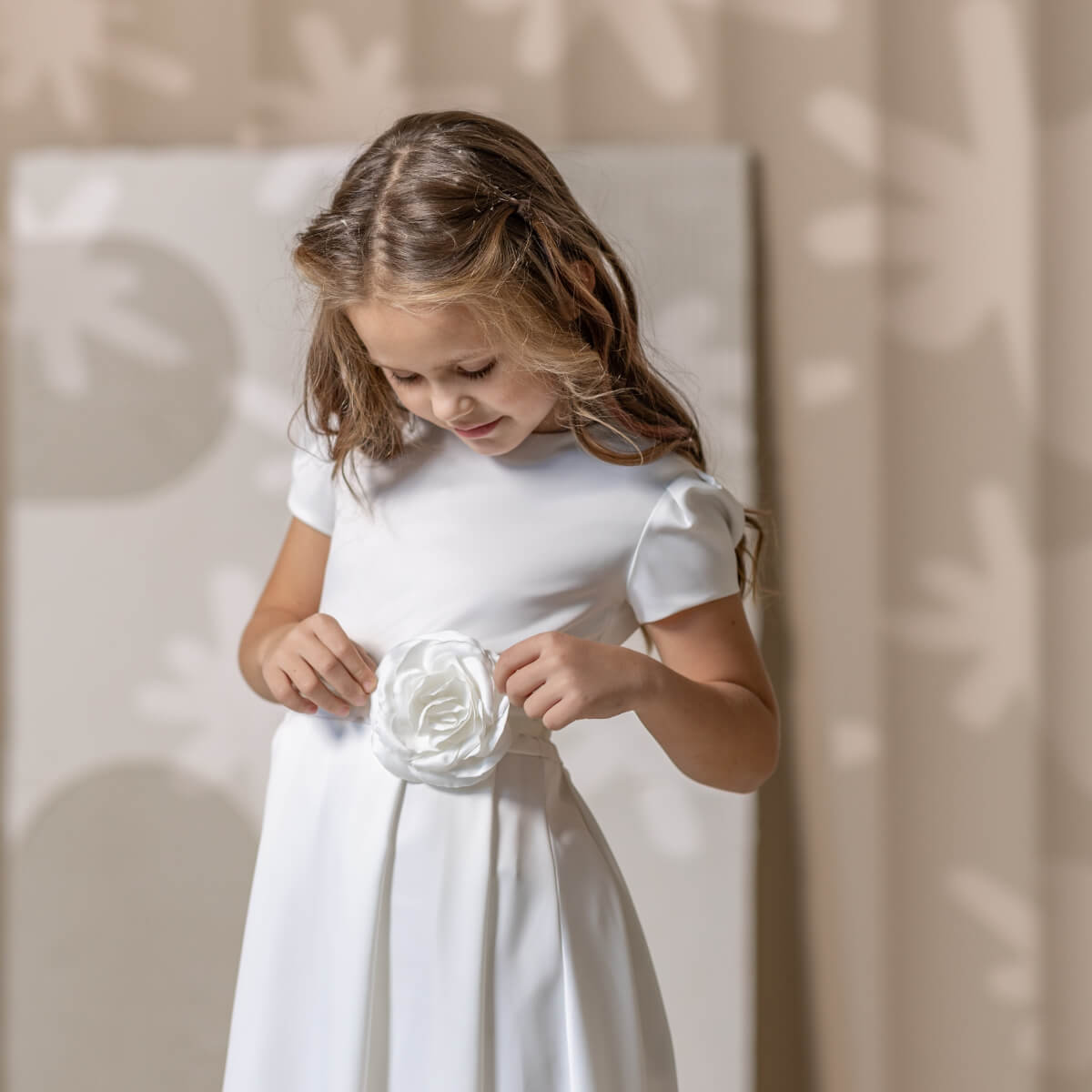 Young girl in a white simple communion dress holding a white flower against a neutral background