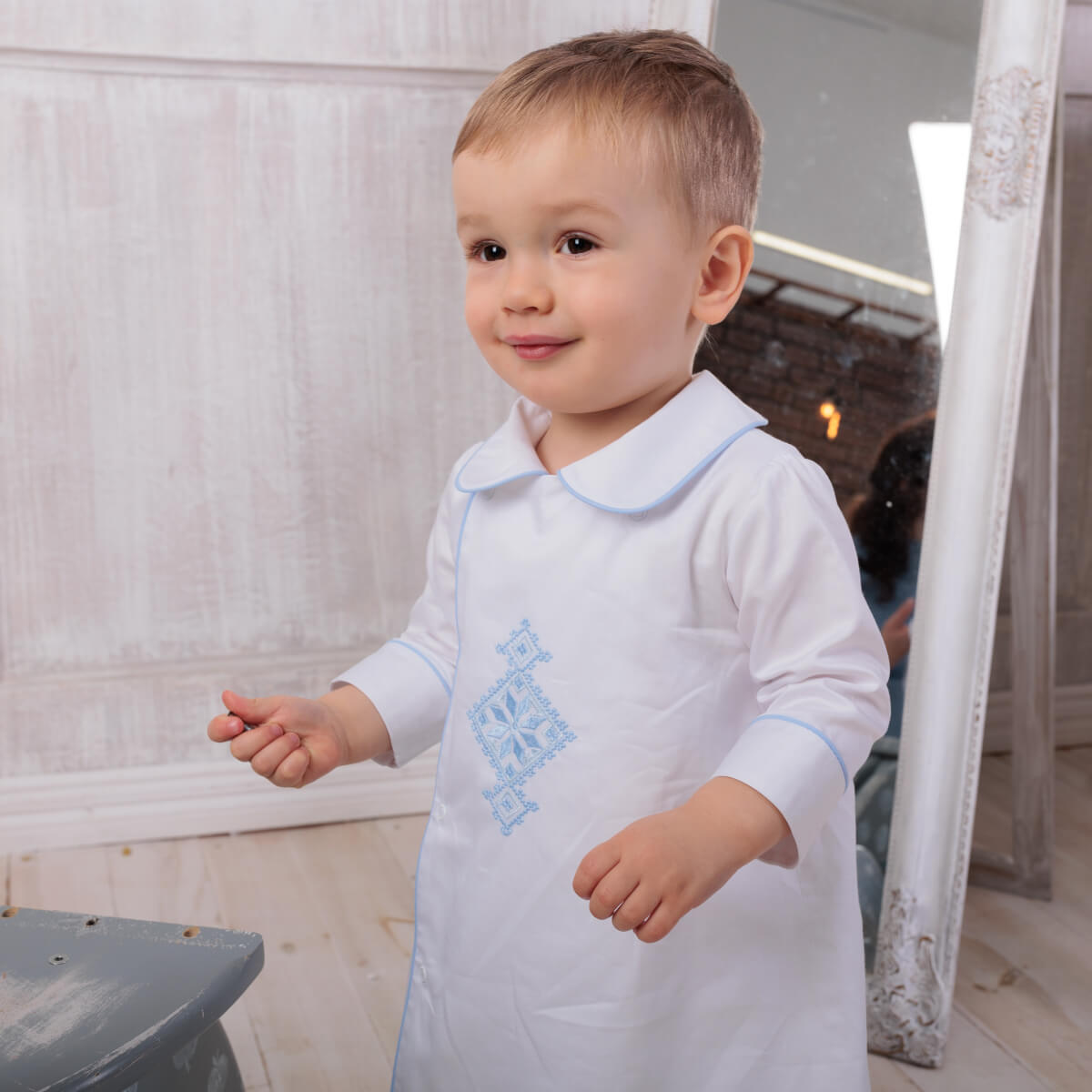 Child wearing a Baptism Gown with blue embroidery in an indoor setting