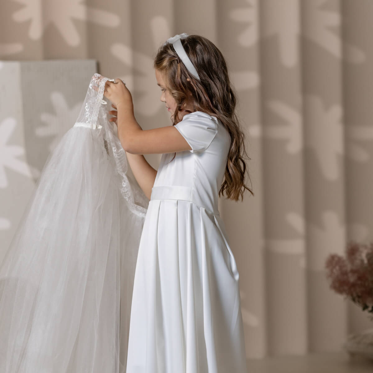 Young girl in a white dress holding a sheer fabric against a neutral background