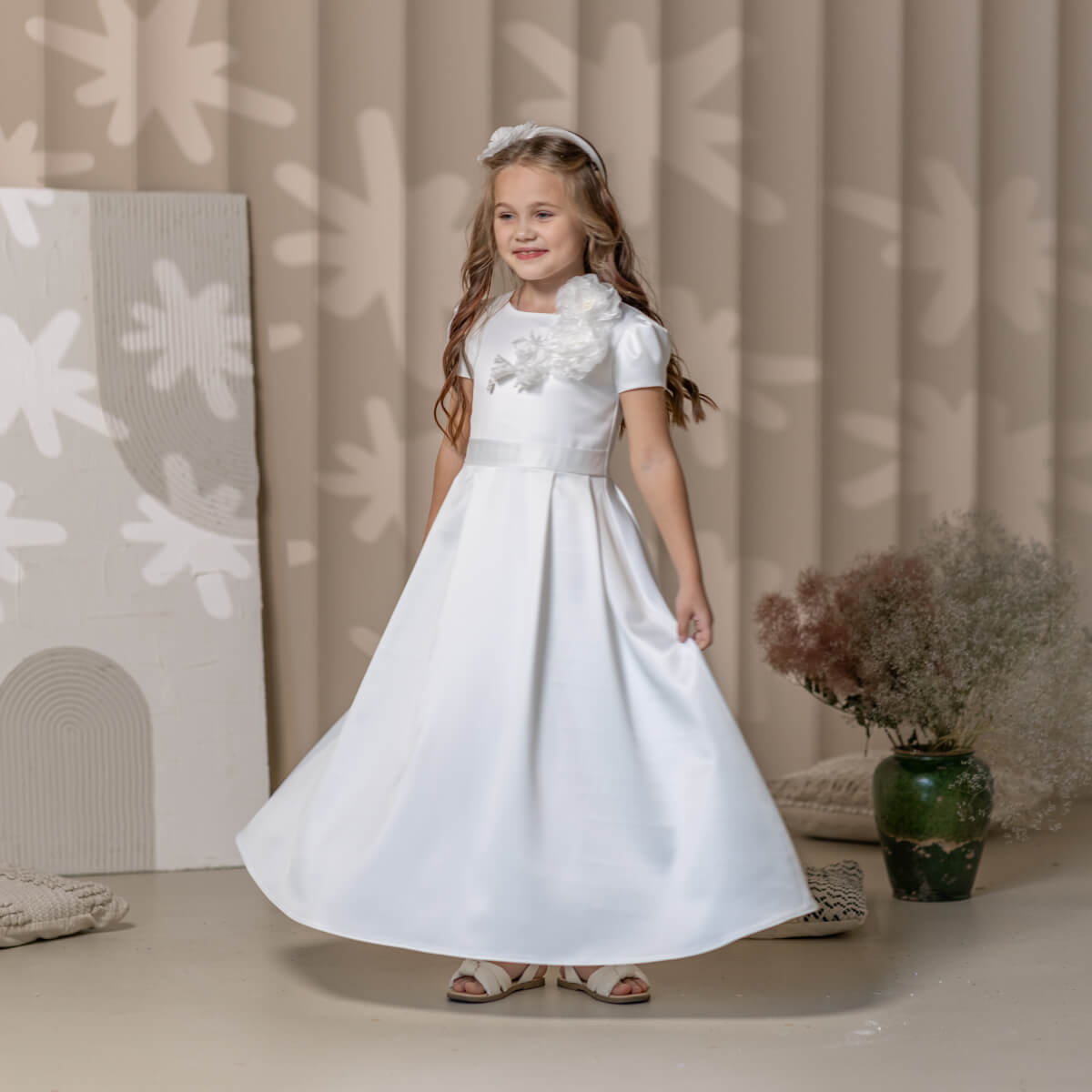 Young girl in a white Catholic Communion Dress standing in a decorated room with floral patterns on the wall.