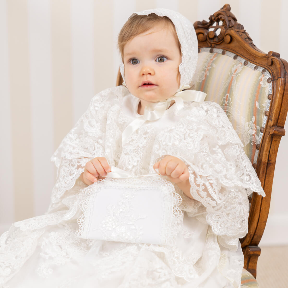 Baby in a white lace outfit sitting on a wooden chair and holding Baptism Keepsake Box.