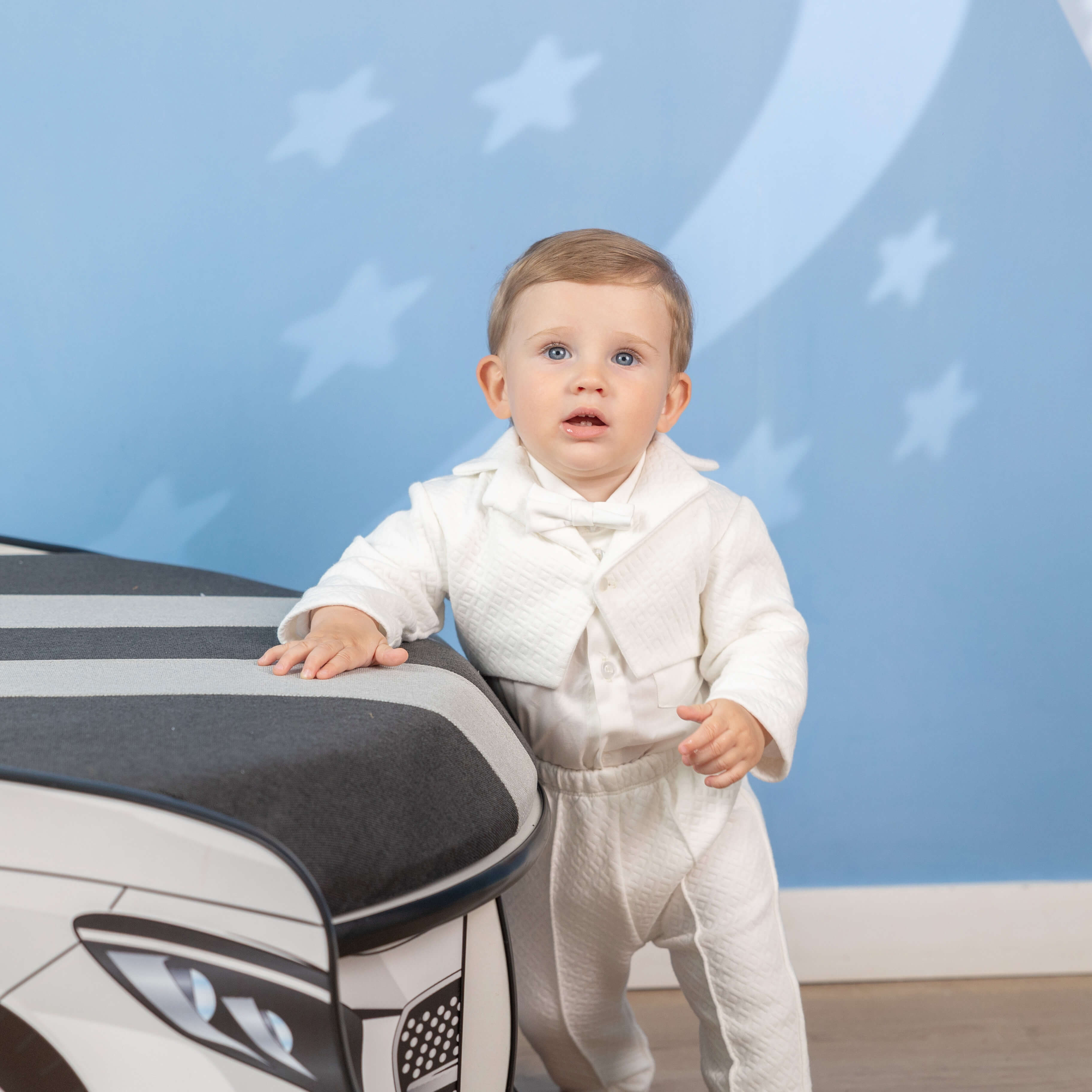 Child in a Baby Boy Baptism Suit – t standing next to a toy car with a blue wall and star decorations in the background