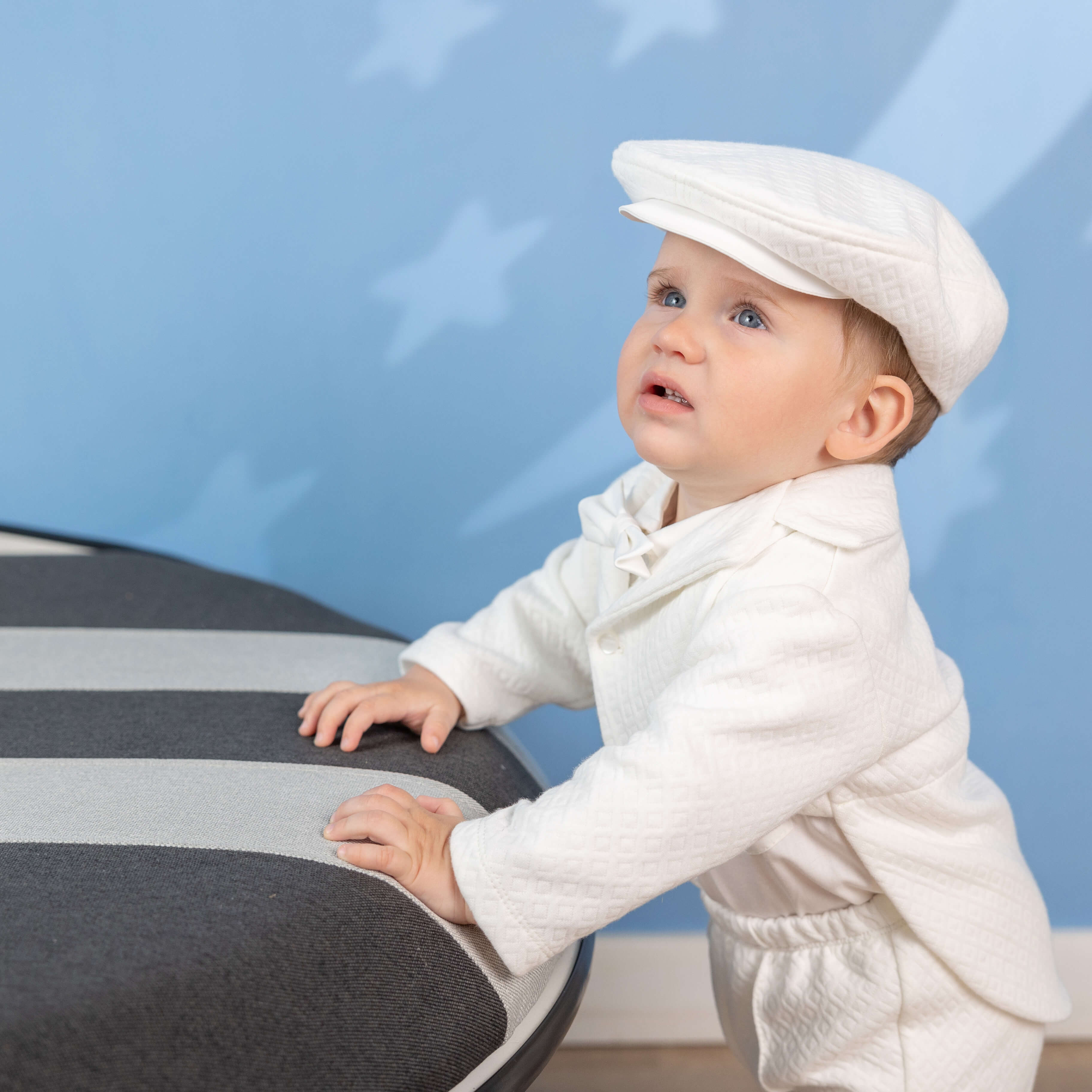 Baby in a Baby Boy Baptism Suit – and cap playing with a toy piano against a blue background