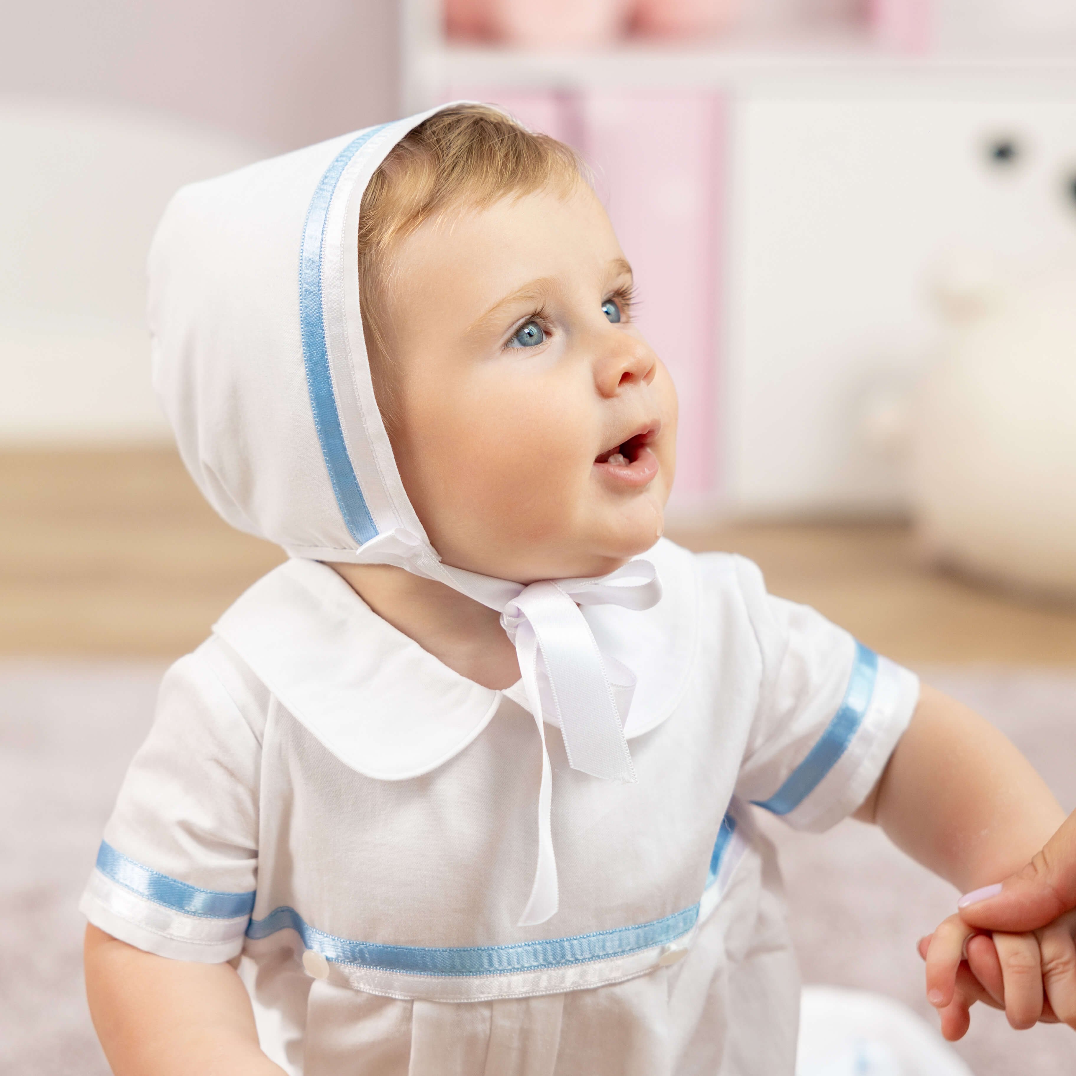 Baby wearing a white dress with blue trim and a bonnet, sitting on a couch.