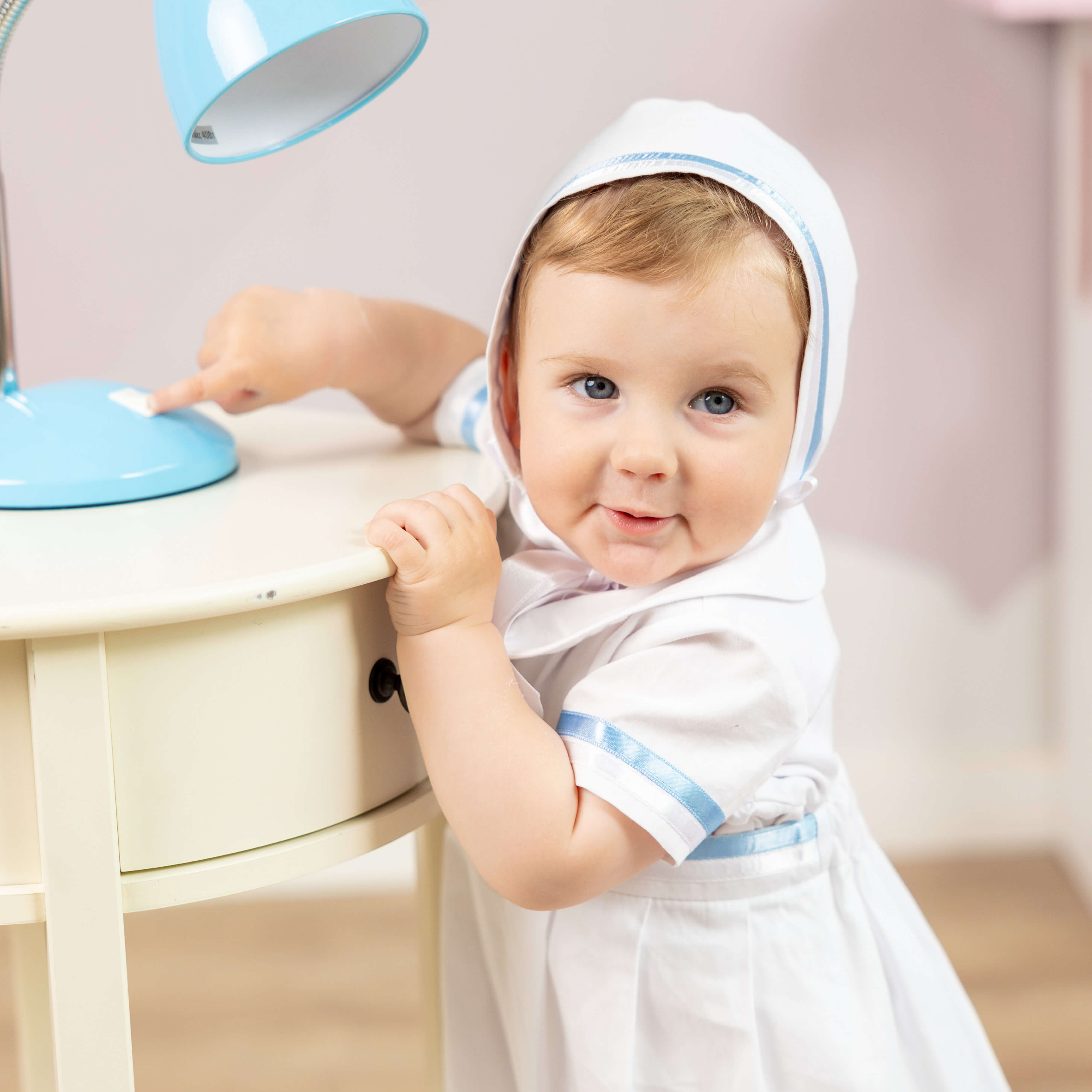 Baby in a Convertible Baptism Gown & Romper. with blue trim sitting at a table.