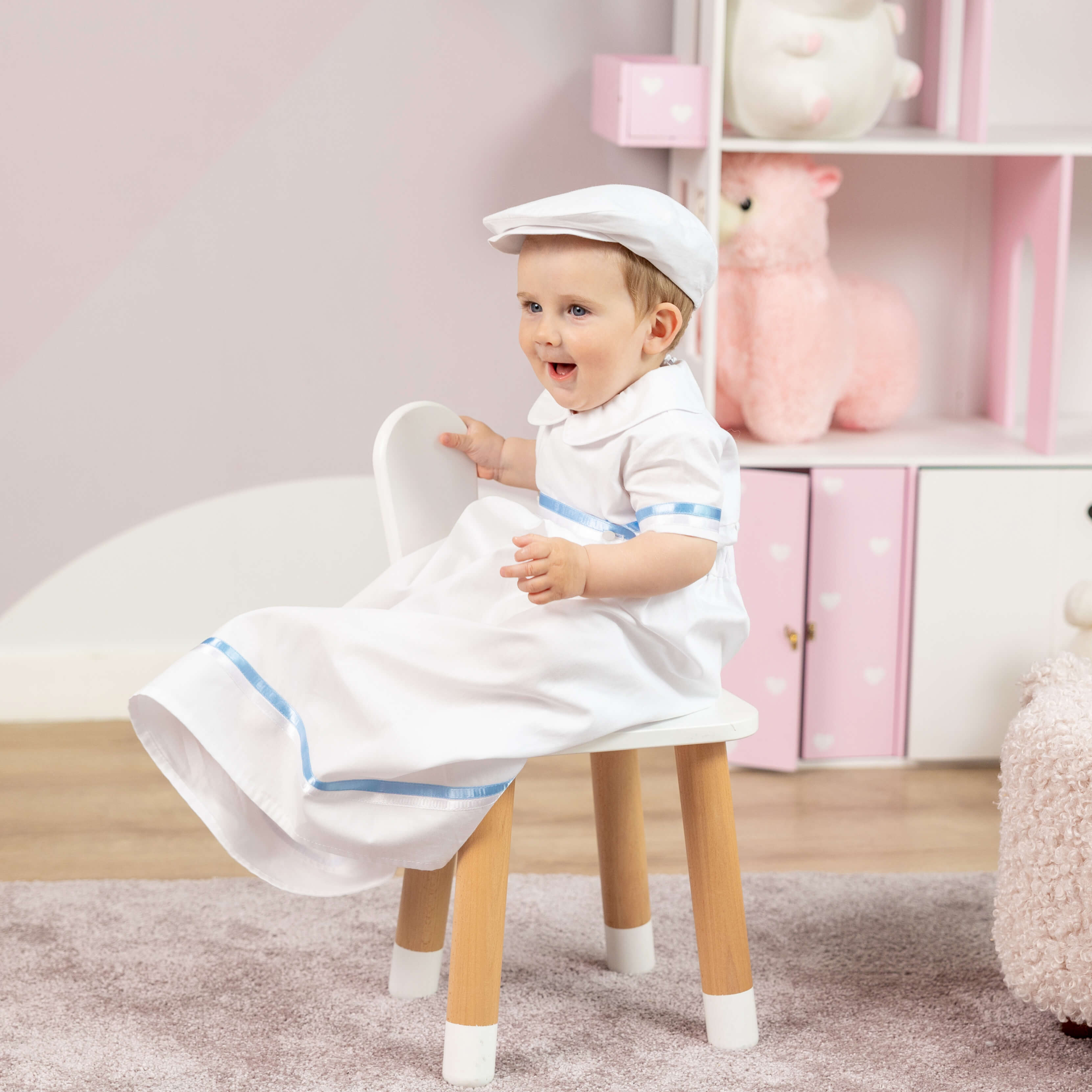 Child in a Convertible Baptism Gown & Rompers with blue trim sitting on a stool in a room with pink decor.