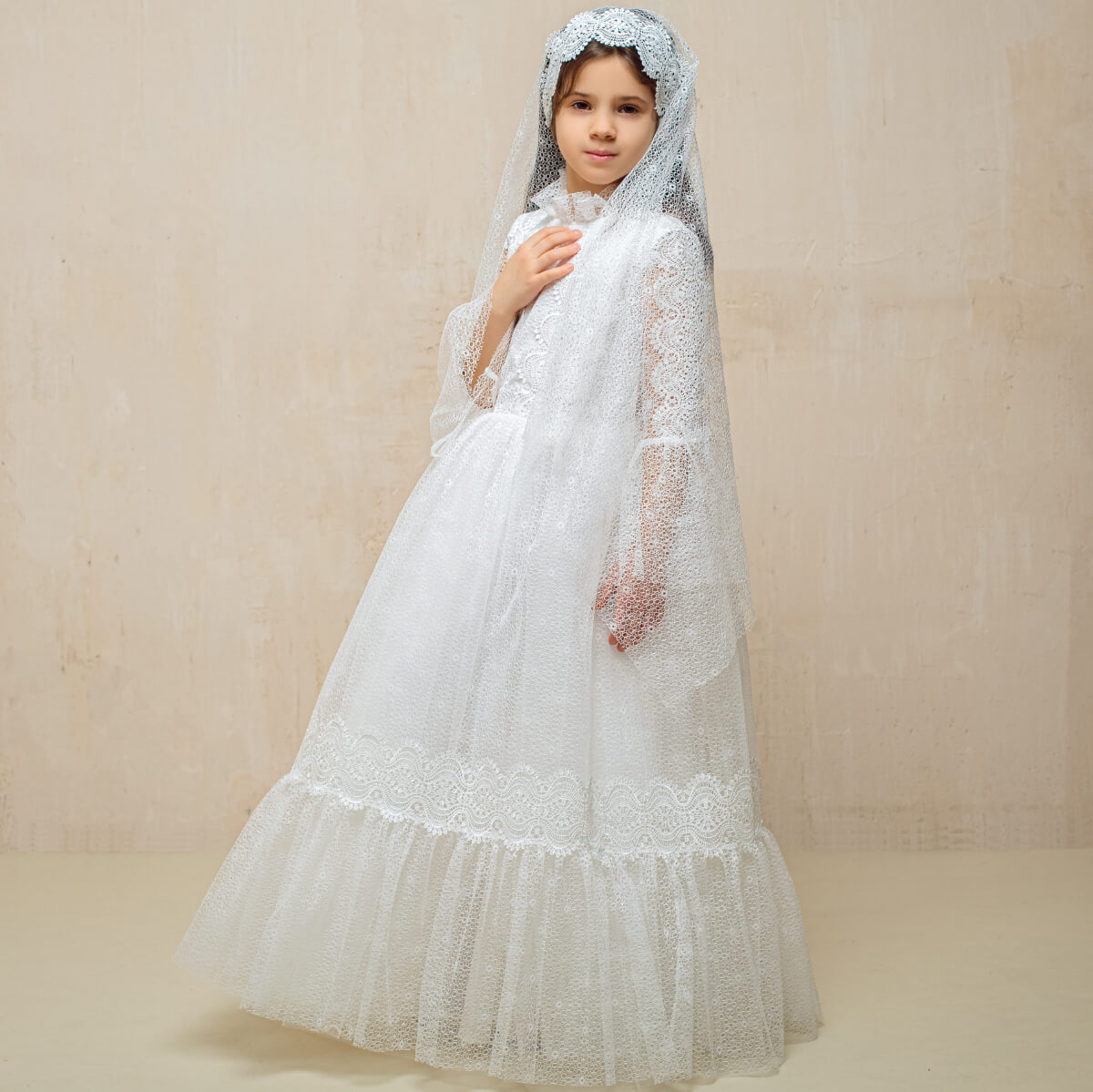Young girl wearing Lace First Communion Dress and head covering against a beige background