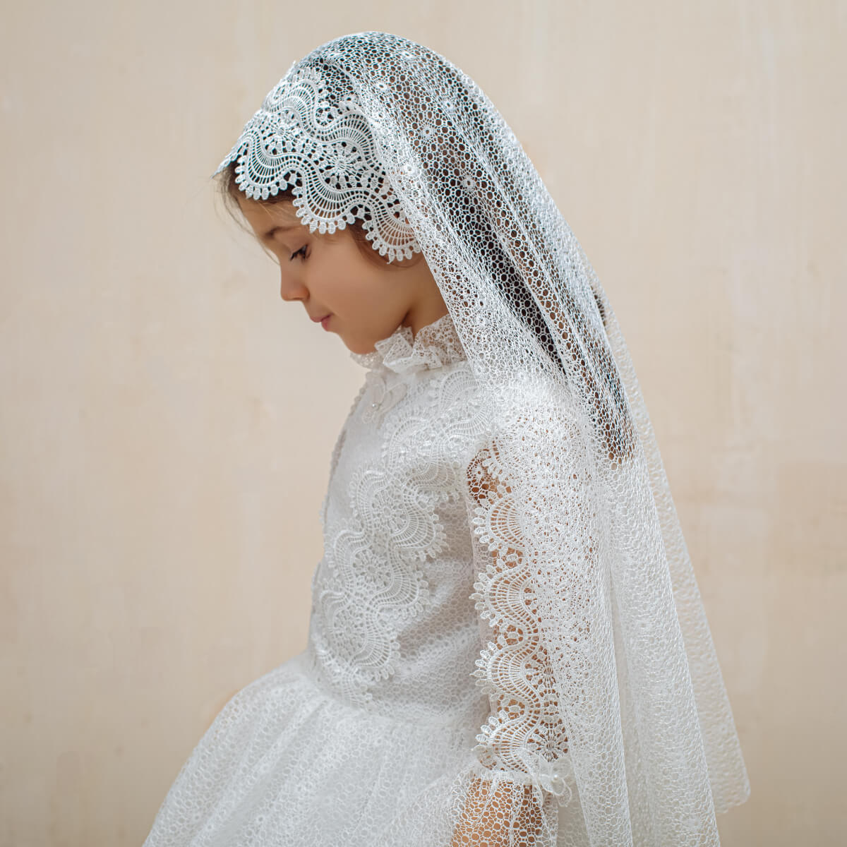 Young girl wearing Lace First Communion Dress and veil against a beige background