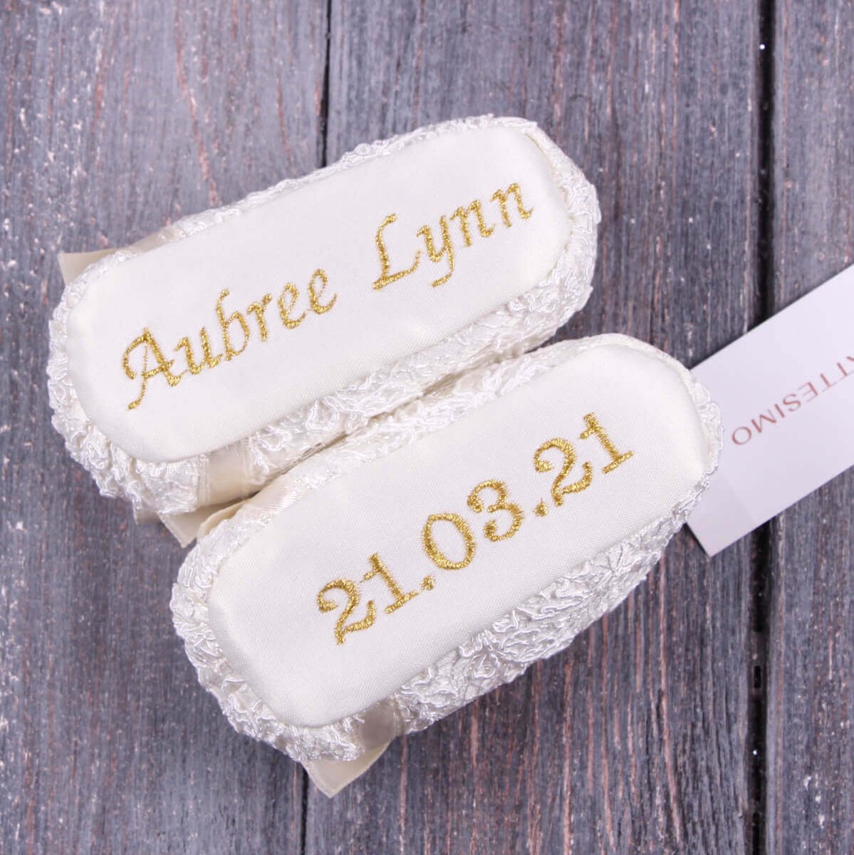 Two white  Christening Booties with gold text on a wooden surface