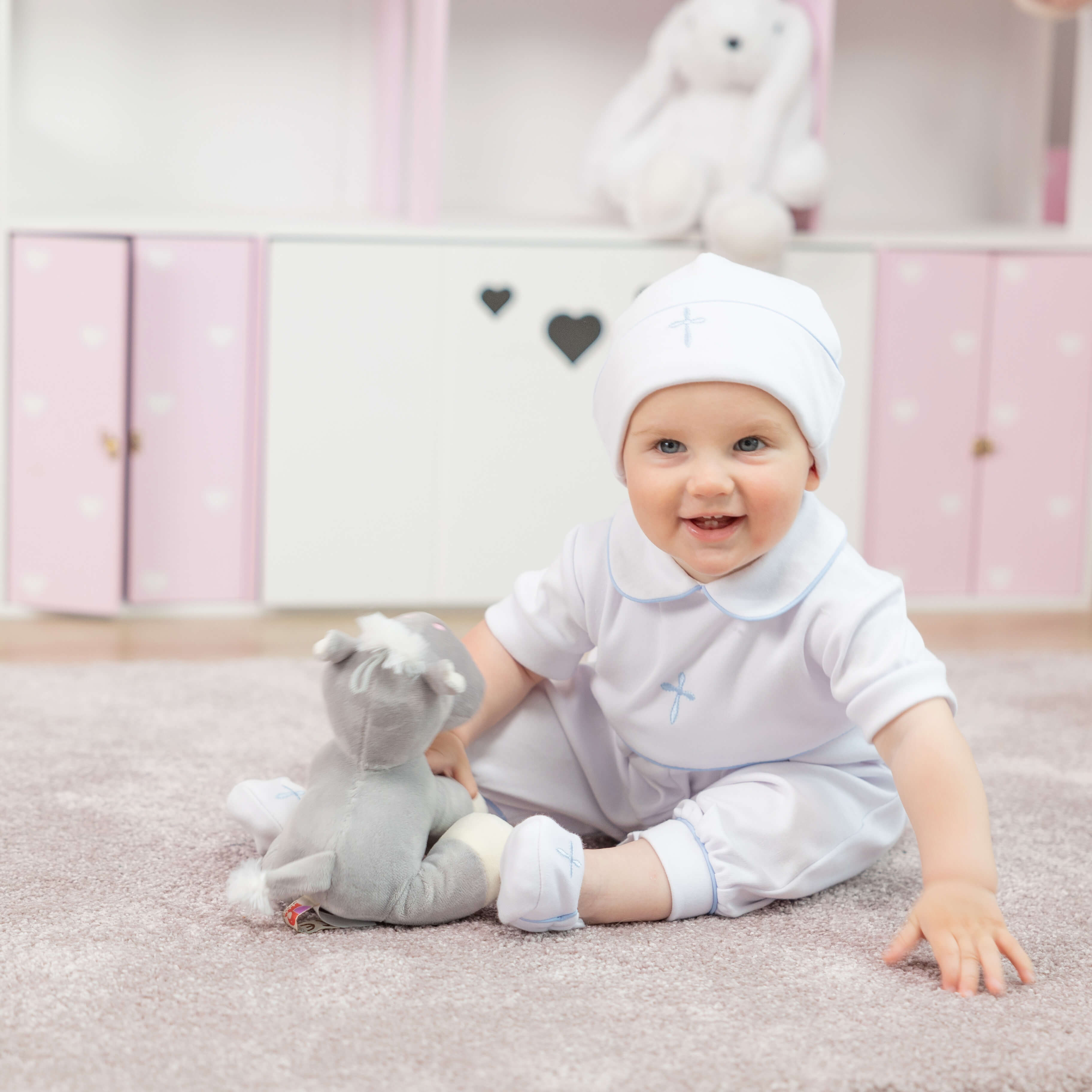 Baby in a  Baptism Outfit for Boys with a hood sitting on a carpeted floor holding a gray toy.