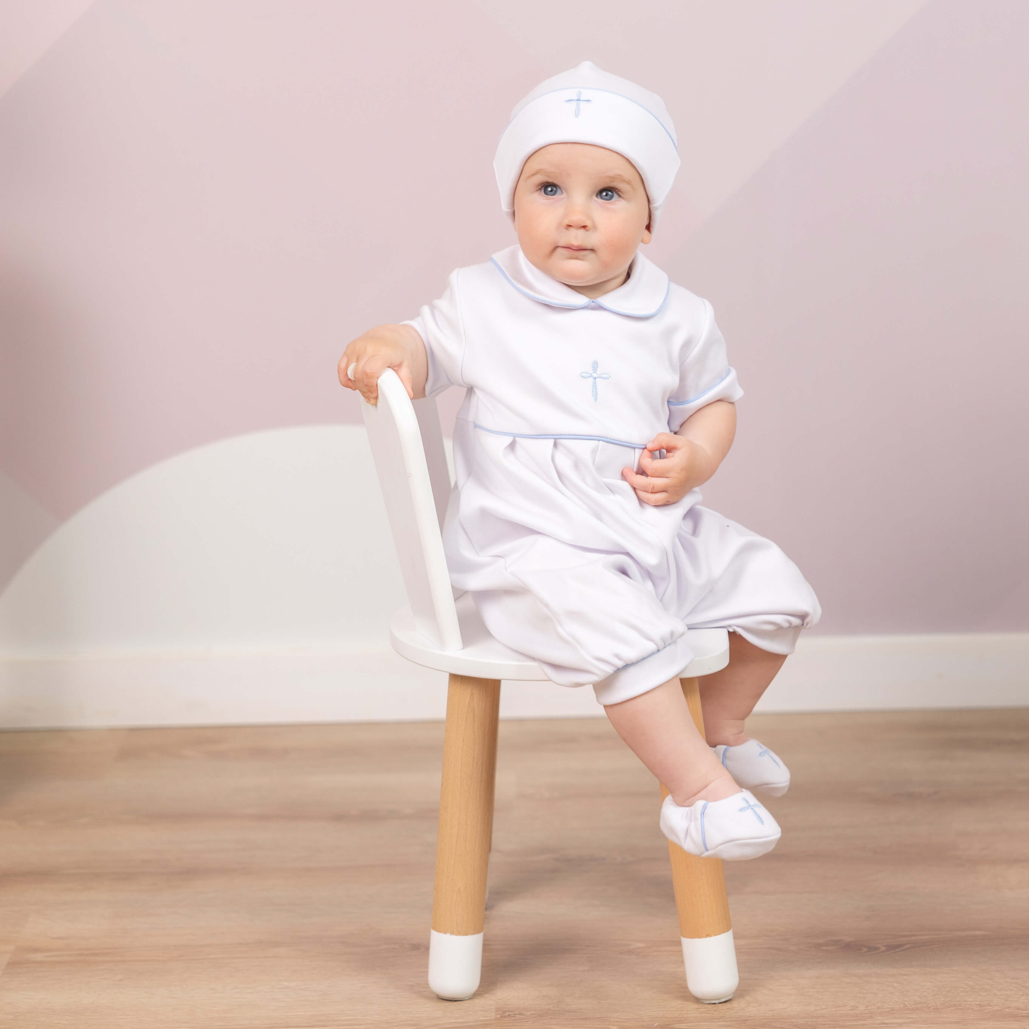 Baby in a  Baptism Outfit for Boys. sitting on a small stool against a light pink background